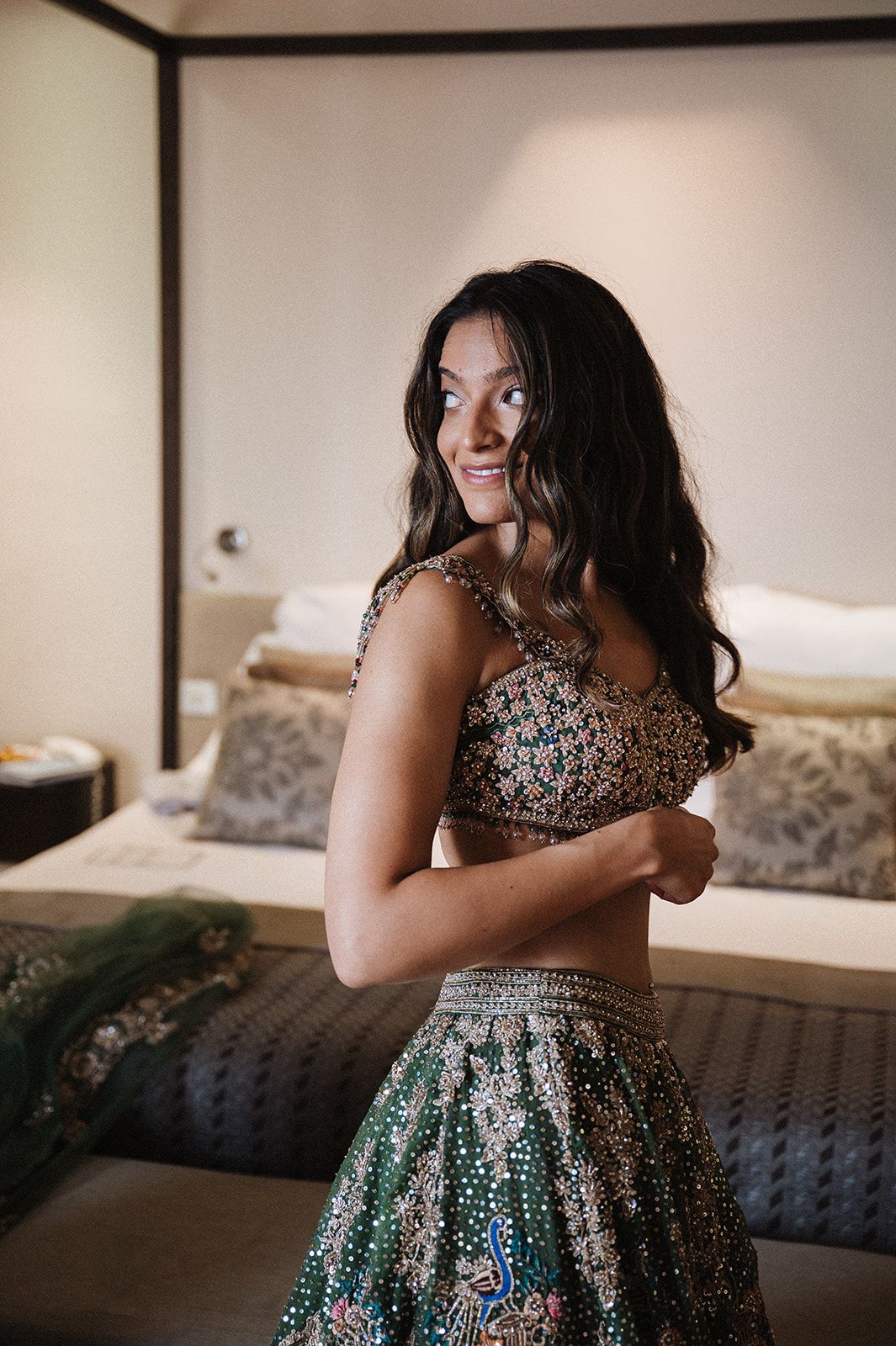 Young woman in ornate green embroidered lehenga choli smiling over shoulder in elegant bedroom setting.