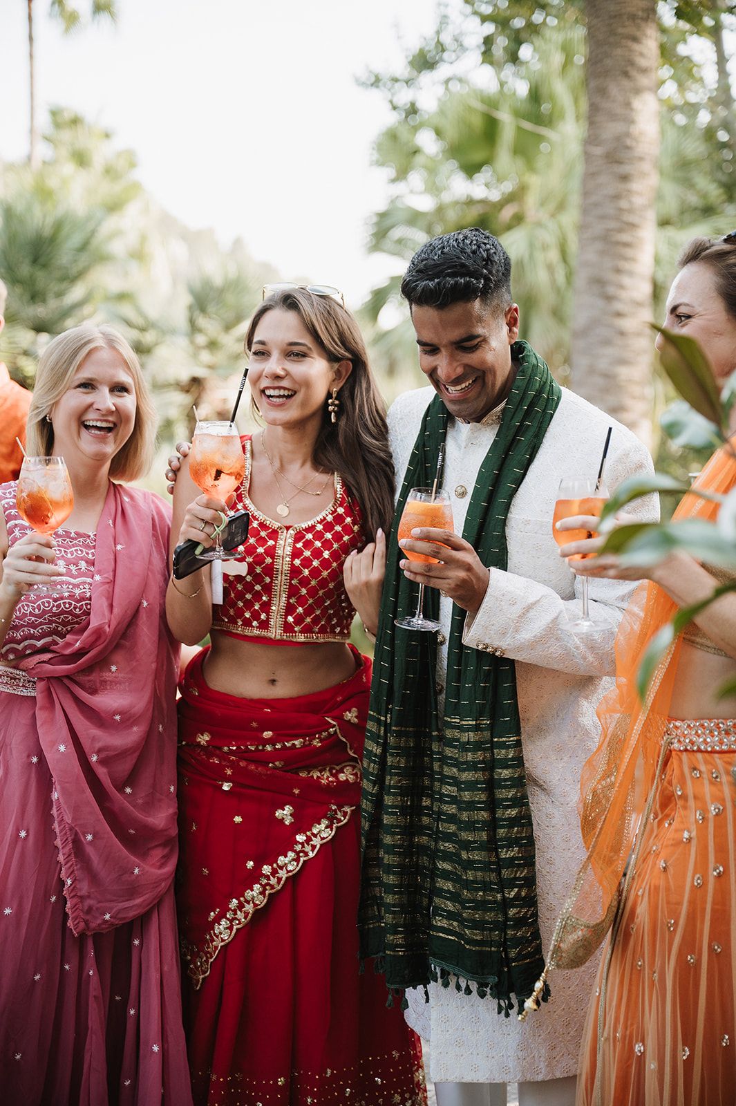Bride in red lehenga and groom in white sherwani with green dupatta celebrate with guests at Indian wedding.