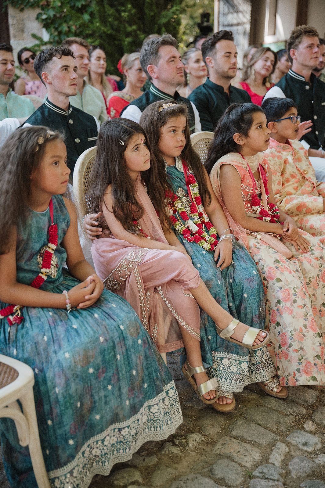 Children in teal Indian outfits with floral garlands seated at an outdoor wedding ceremony.