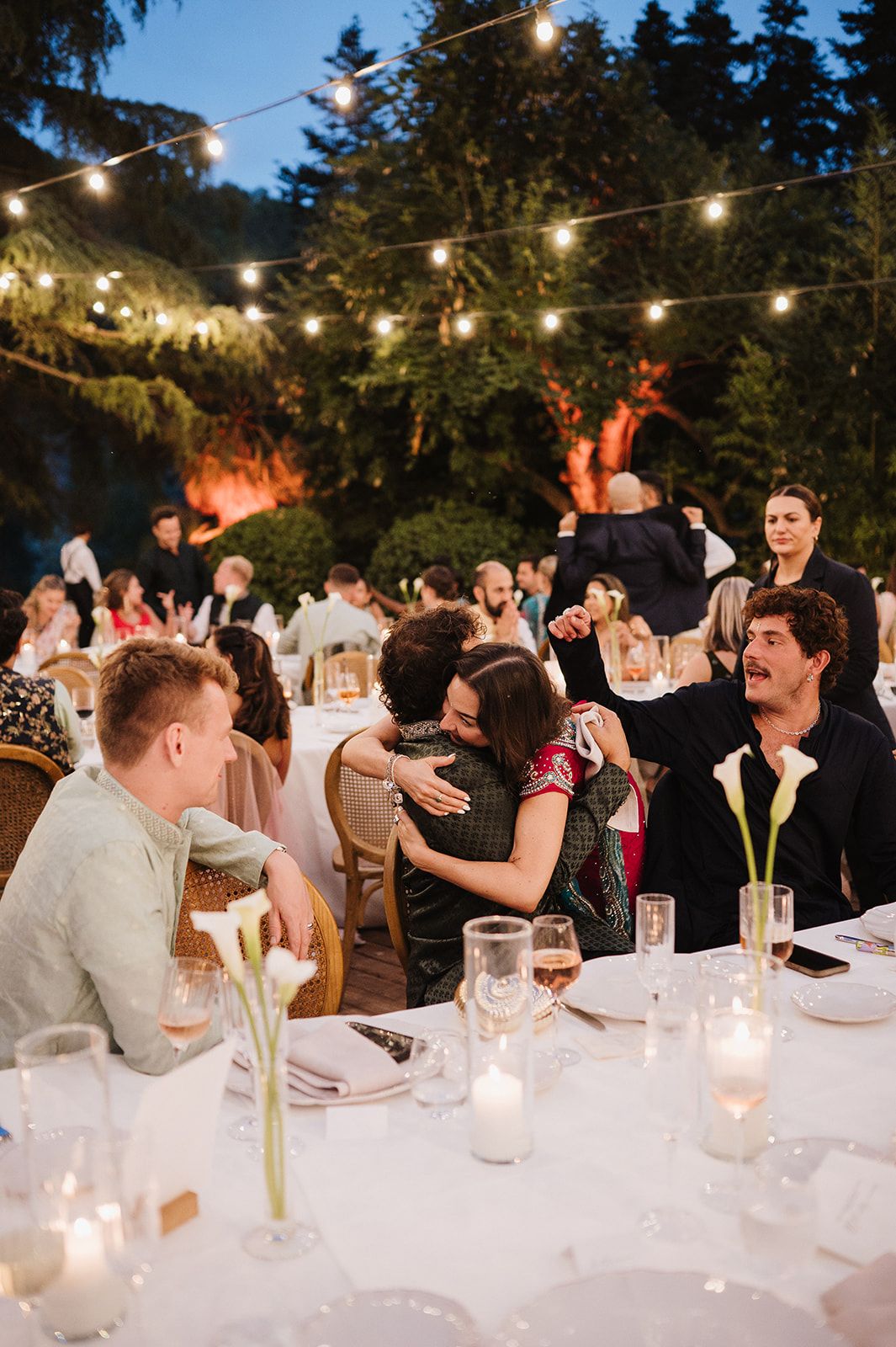 Wedding reception guests embrace and laugh under warm string lights during an outdoor evening dinner.