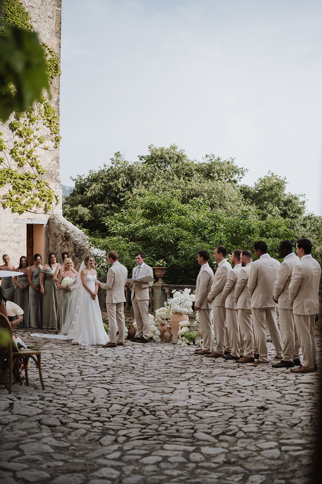Wedding ceremony at a rustic cobblestone venue with bridesmaids and groomsmen in neutral tones.