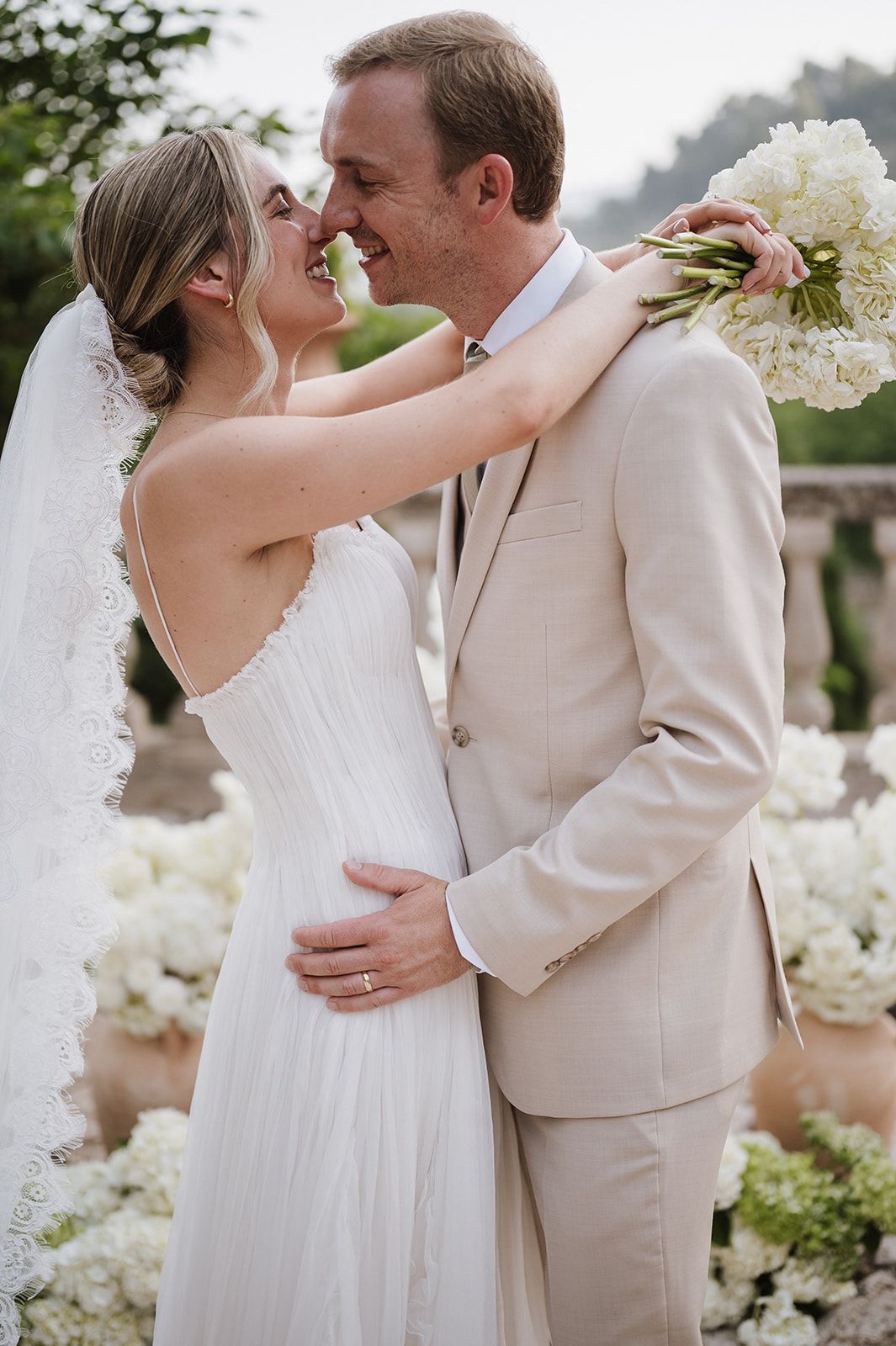 Smiling bride in lace veil embraces groom in beige suit holding white hydrangea bouquet outdoors.