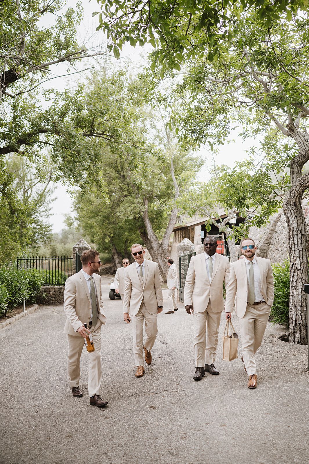 Four groomsmen in matching beige suits walk confidently down a tree-lined outdoor pathway.