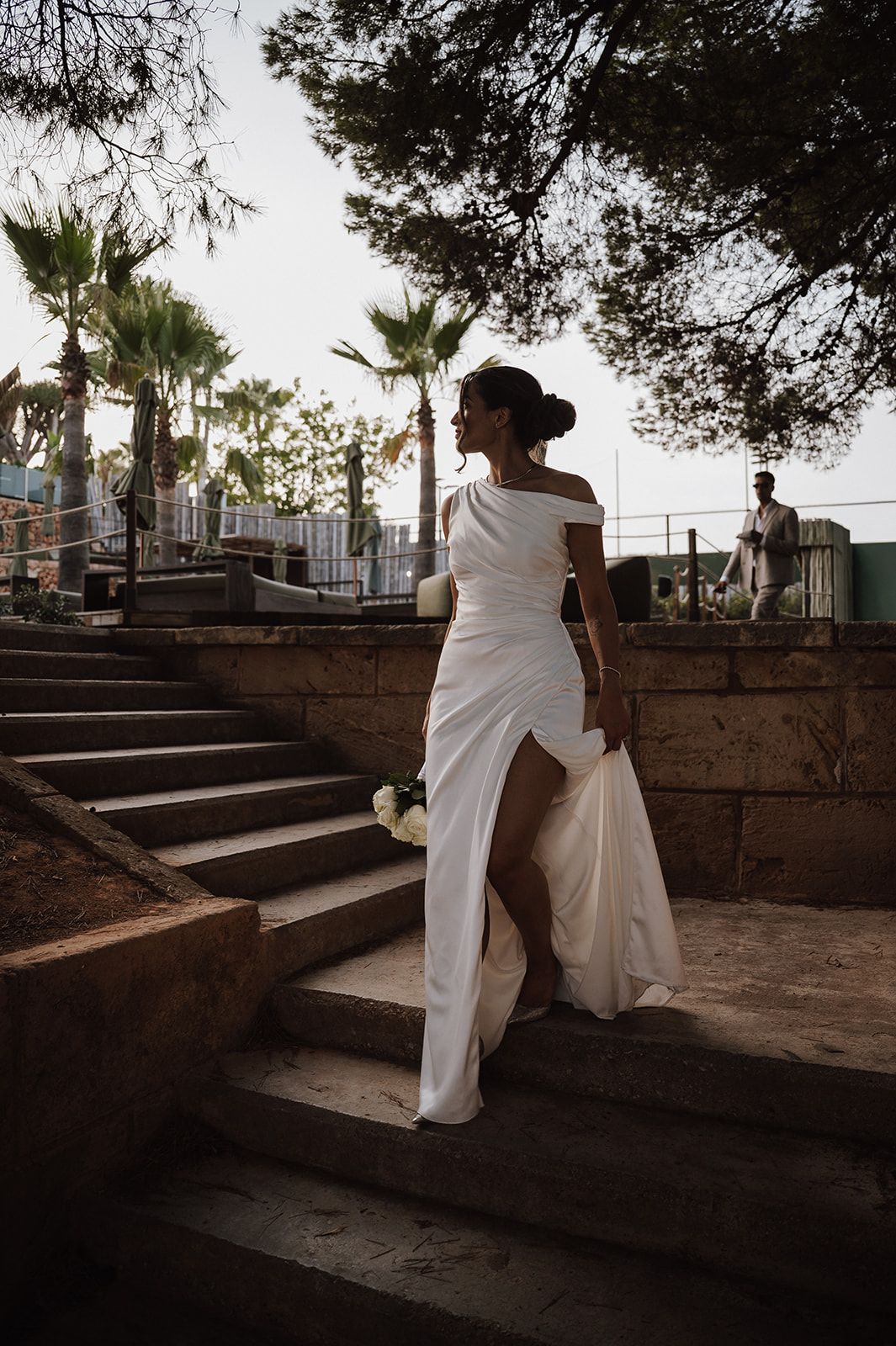 Bride in off-shoulder white gown with slit poses on stone steps at sunset.