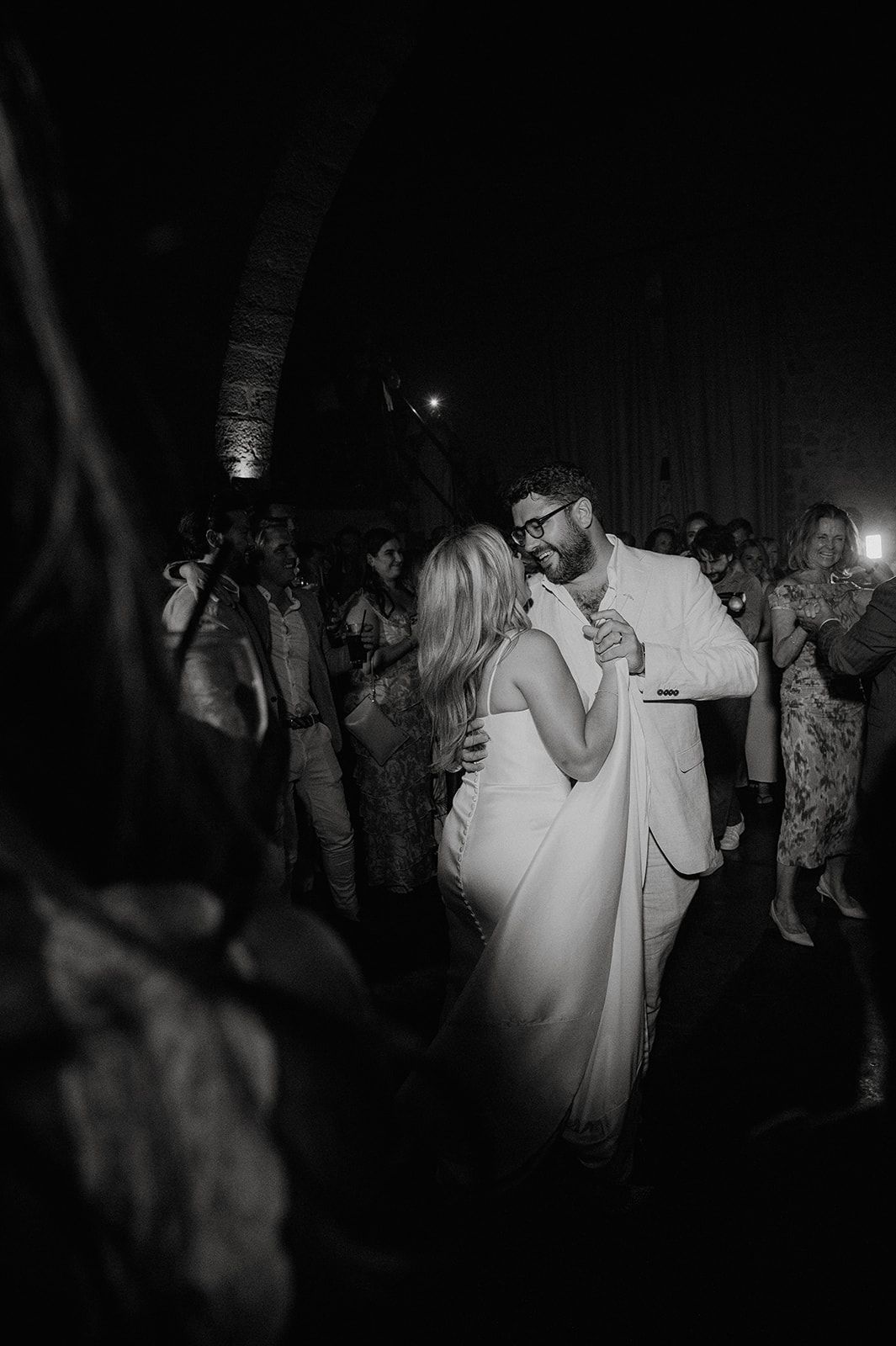 Bride and groom share their first dance surrounded by guests in a romantic black and white wedding photo.