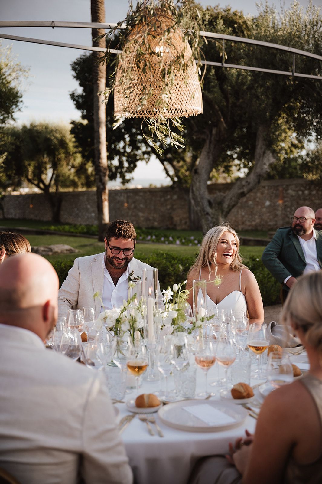 Bride and groom laugh joyfully with guests at outdoor wedding reception dinner under woven chandelier.