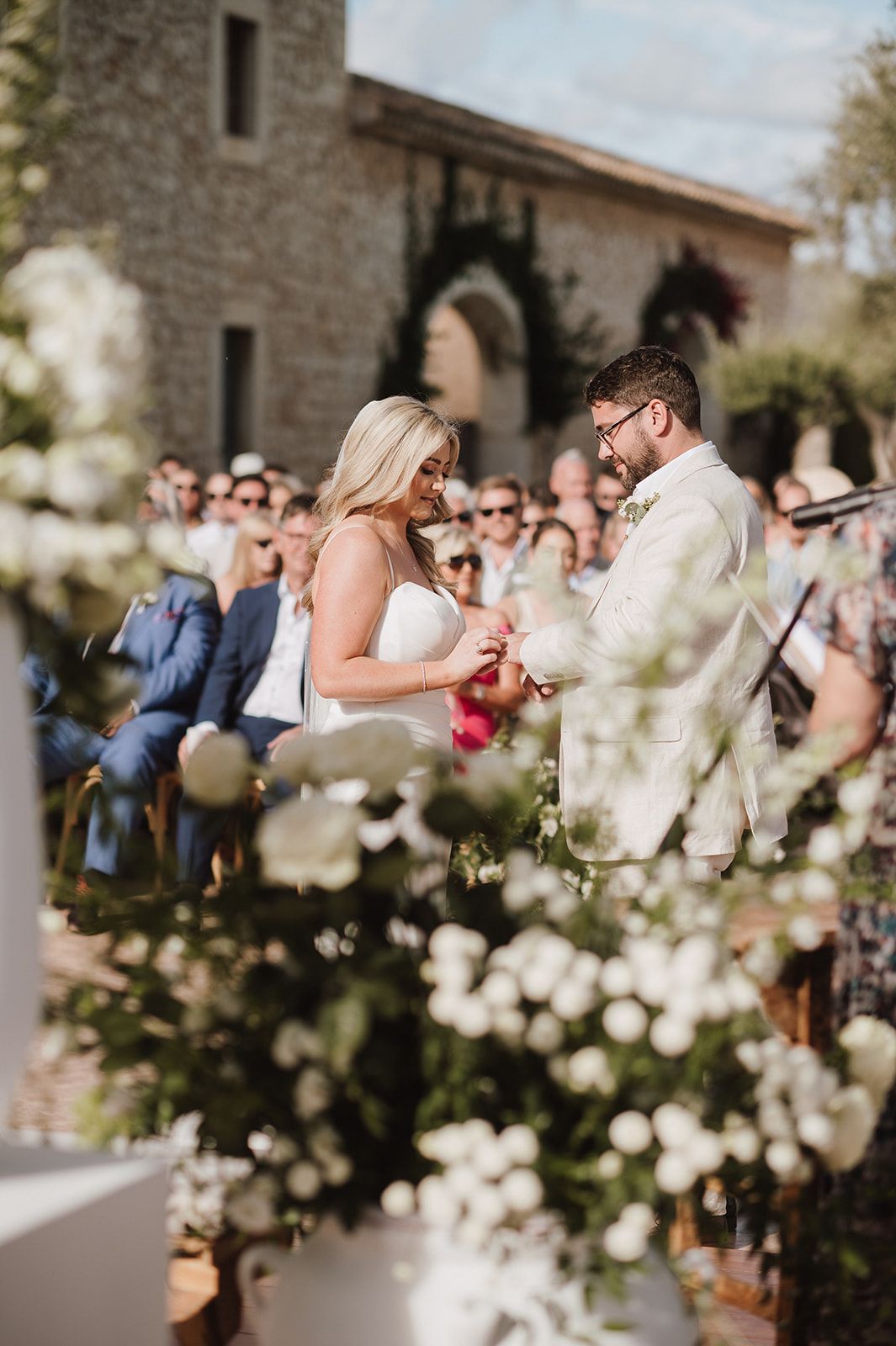 Bride and groom exchange rings during outdoor wedding ceremony surrounded by white floral arrangements.
