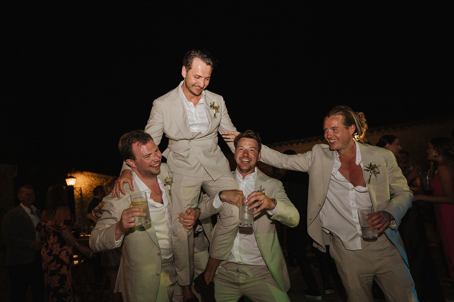 Groom lifted on shoulders by groomsmen in matching beige suits at outdoor wedding party.