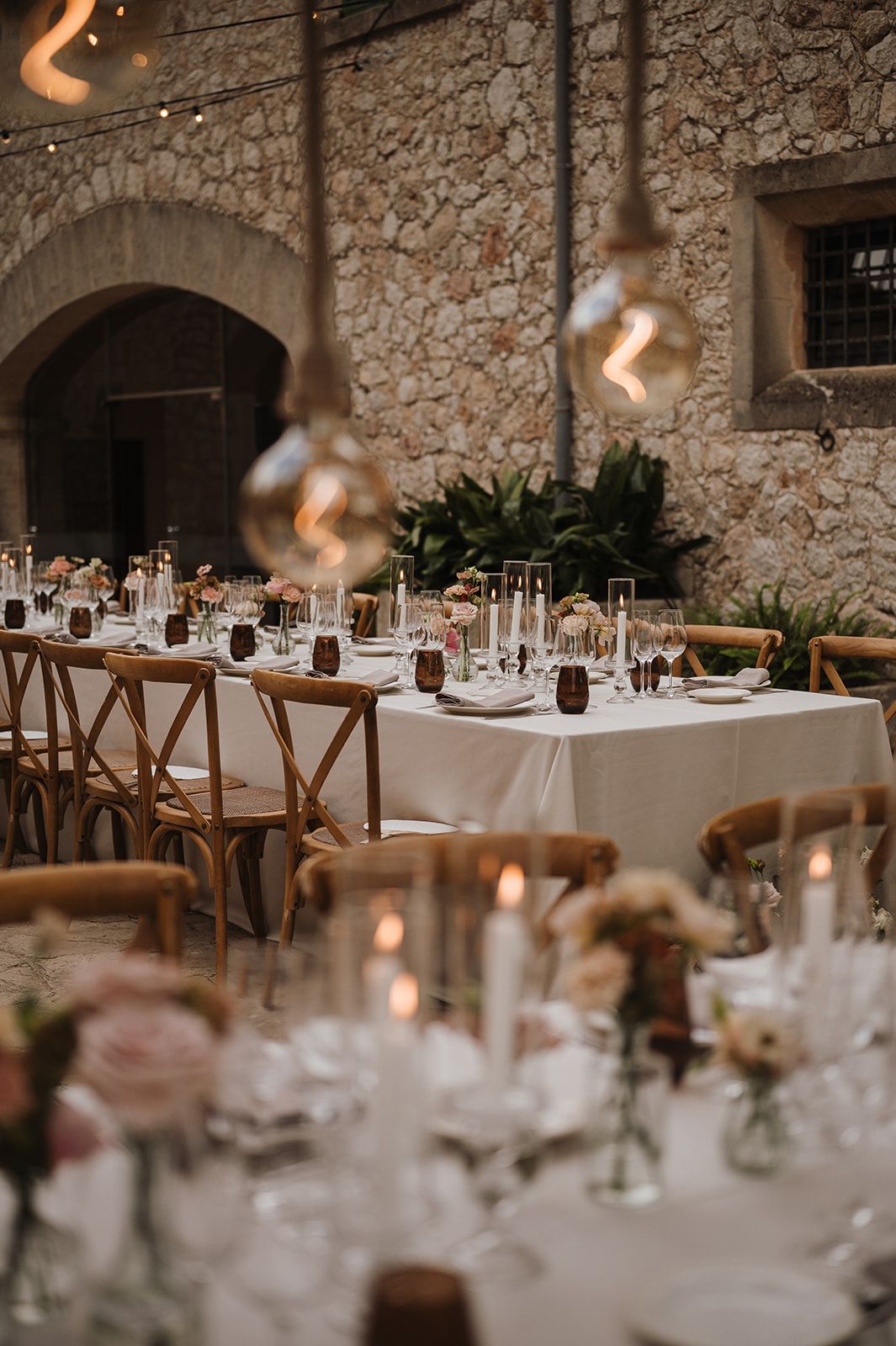 Candlelit outdoor wedding reception tables with globe pendant lights in a rustic stone courtyard venue.