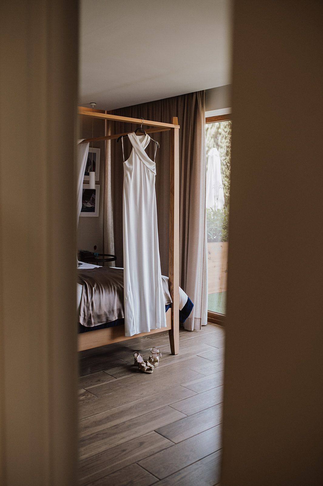 White halter wedding dress hanging on wooden rack in sunlit modern hotel room.