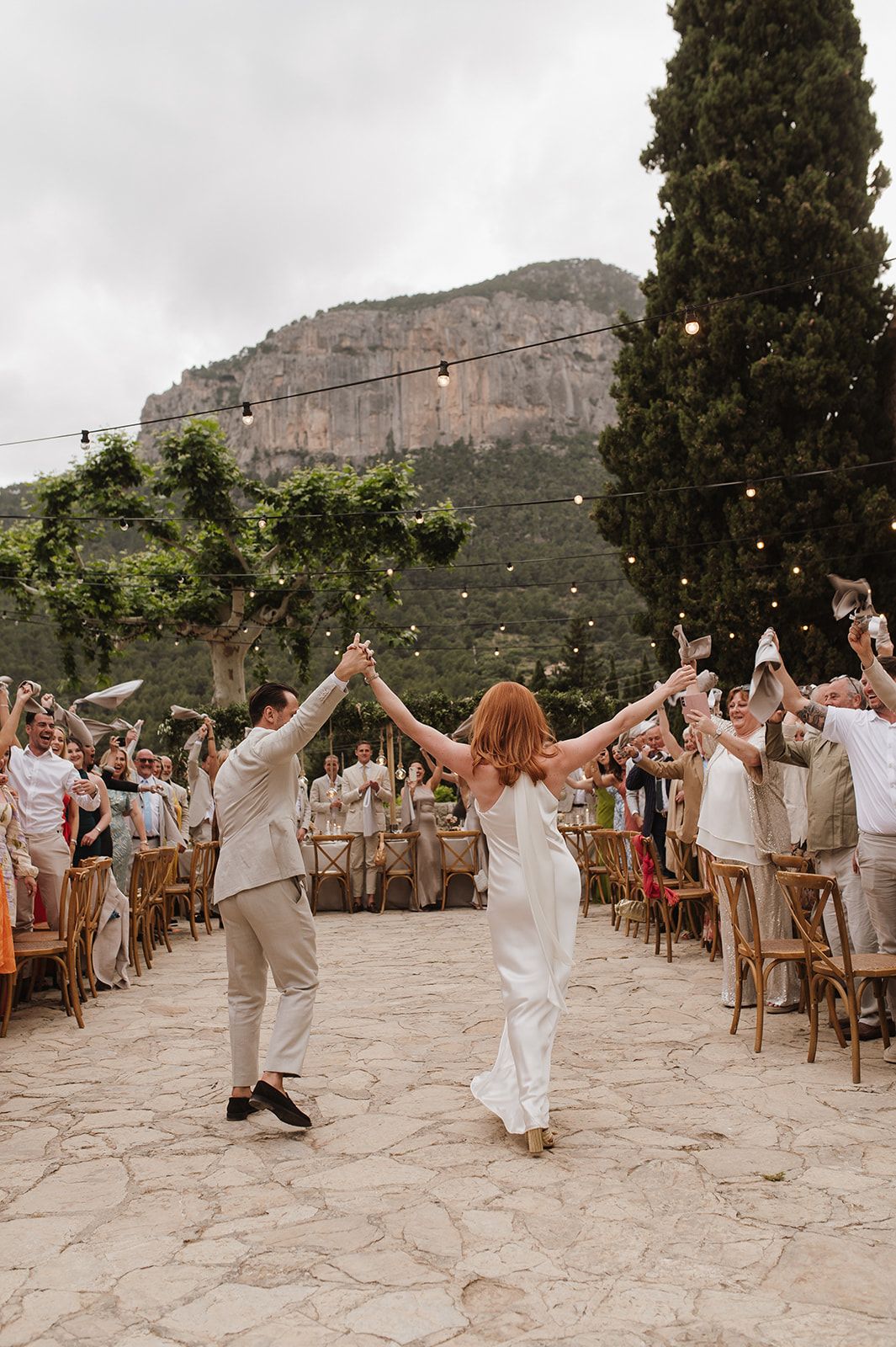 Bride and groom walk hand-in-hand through cheering guests at outdoor mountain wedding reception.