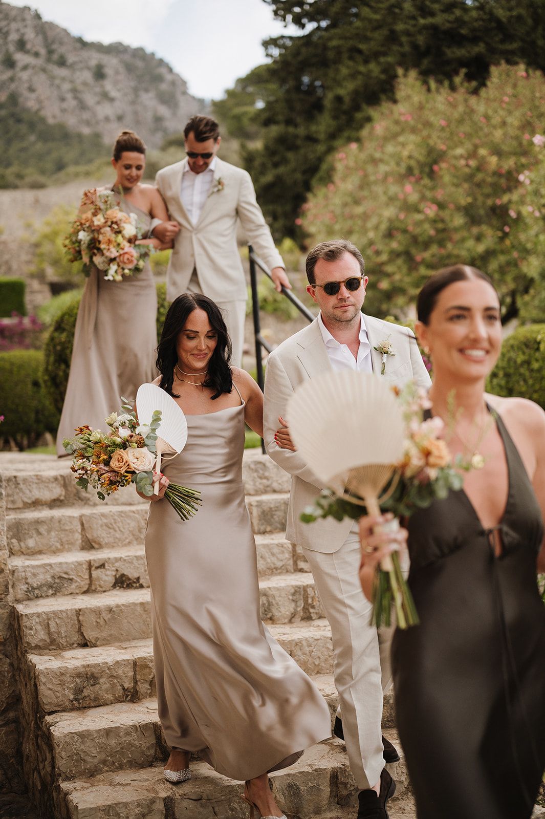 Wedding party descends stone steps carrying bouquets at scenic outdoor ceremony with mountain backdrop.