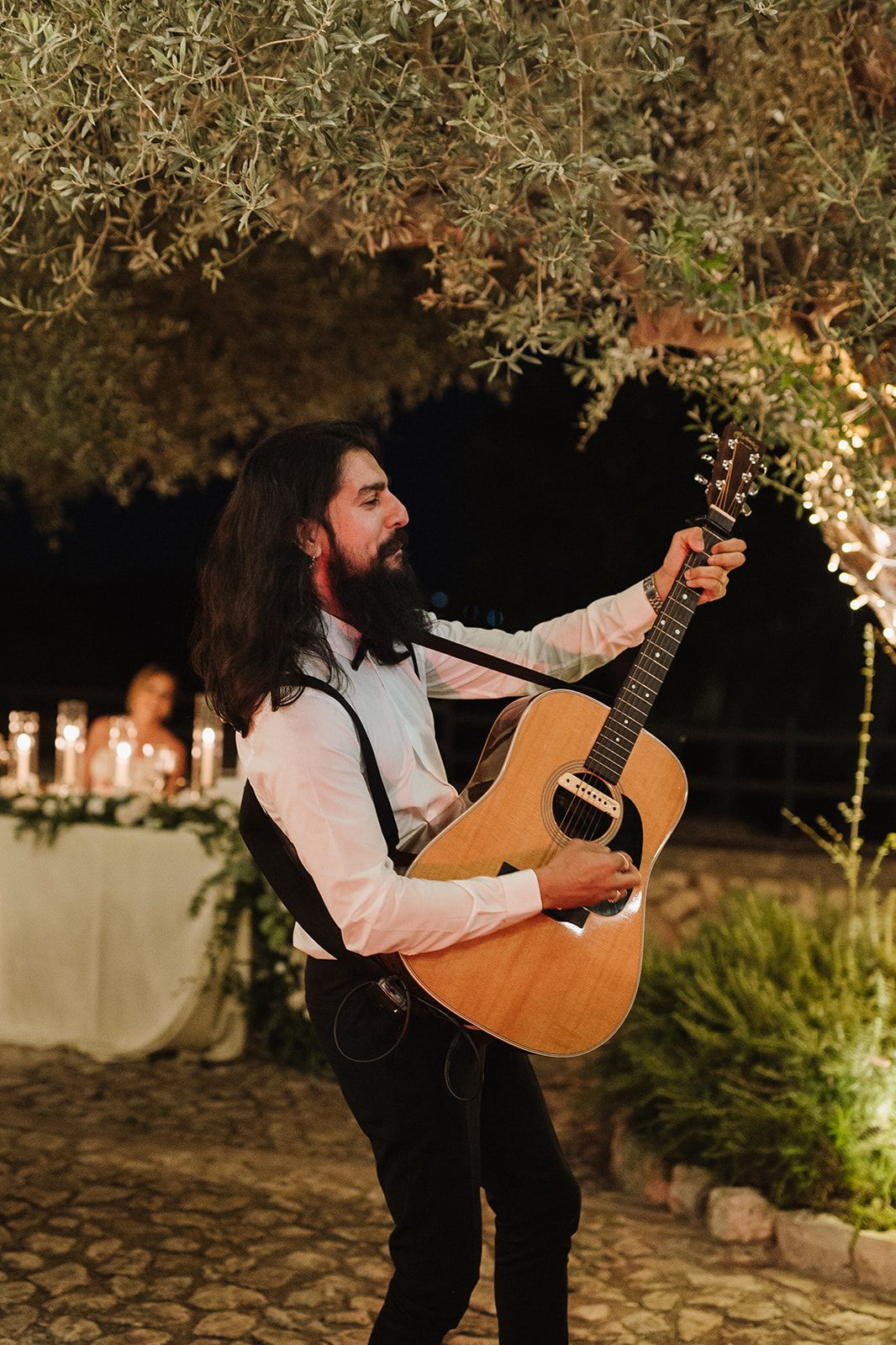 Long-haired guitarist with beard performs under olive tree at outdoor wedding reception at night.