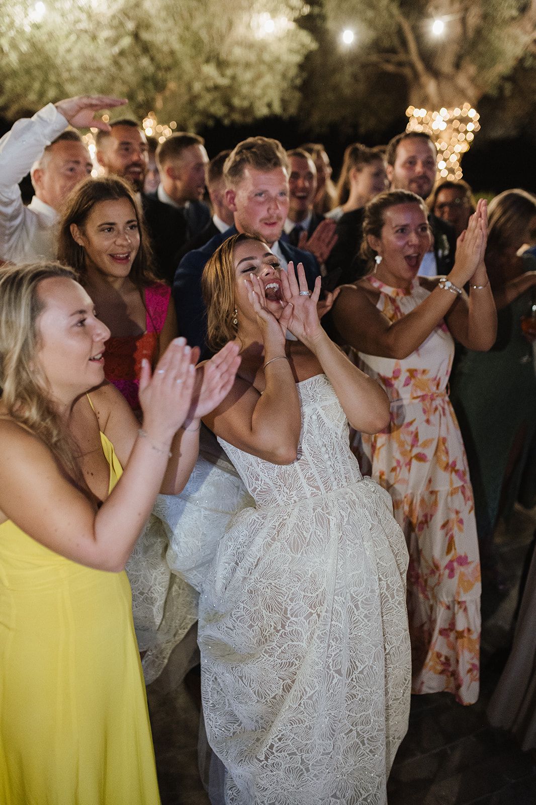 Excited bride in lace gown cheering with wedding guests clapping and celebrating at an outdoor evening reception.