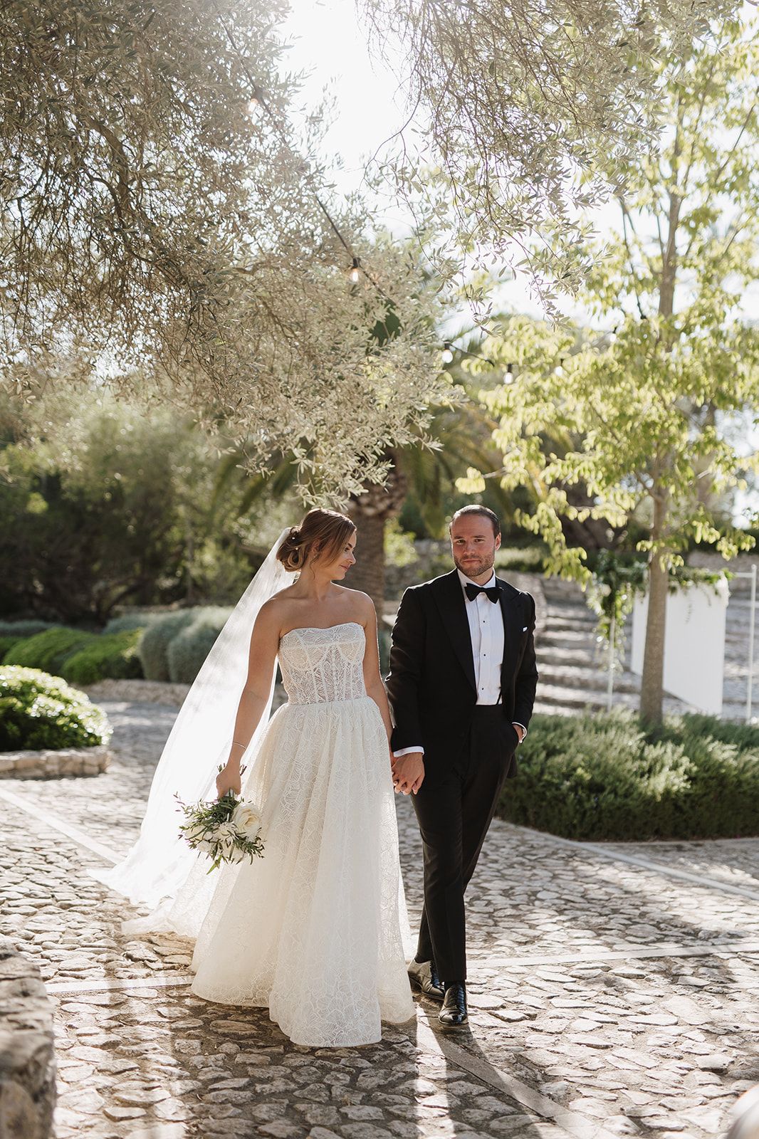 Bride in strapless lace gown and groom in black tuxedo walking hand in hand on cobblestone path.