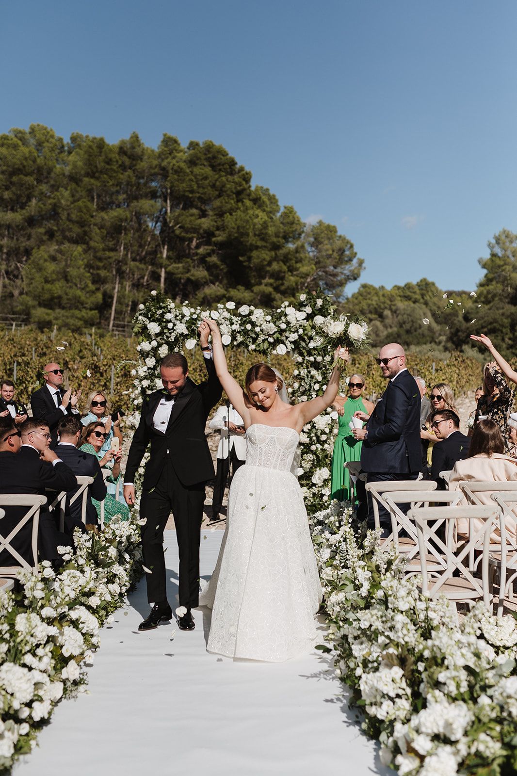 Newlyweds raise hands in celebration walking down white aisle lined with white flowers after ceremony.