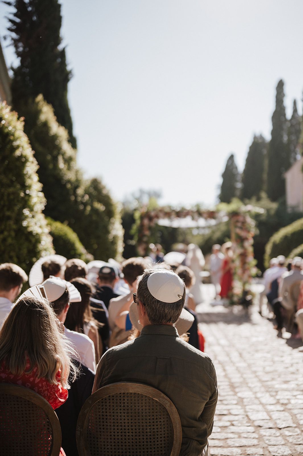 Wedding guests seated outdoors wearing kippahs during Jewish ceremony with floral chuppah ahead.