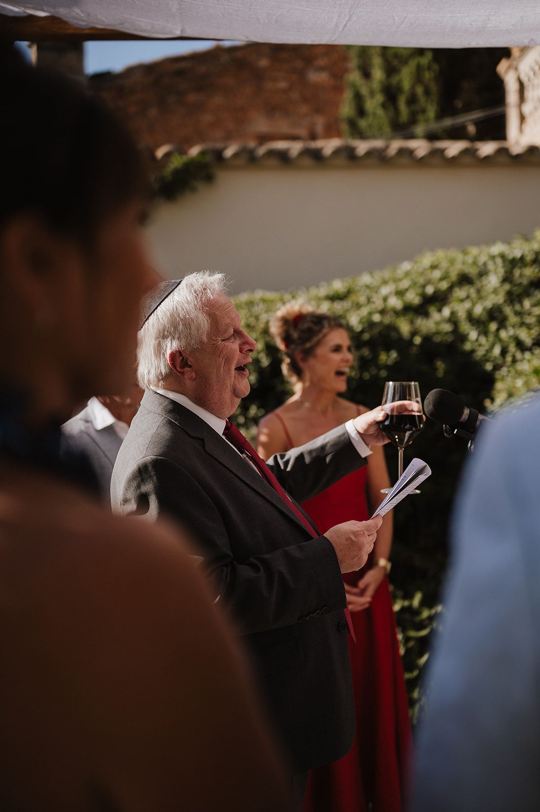 Elderly man in kippah reads at Jewish wedding ceremony while woman in red watches.