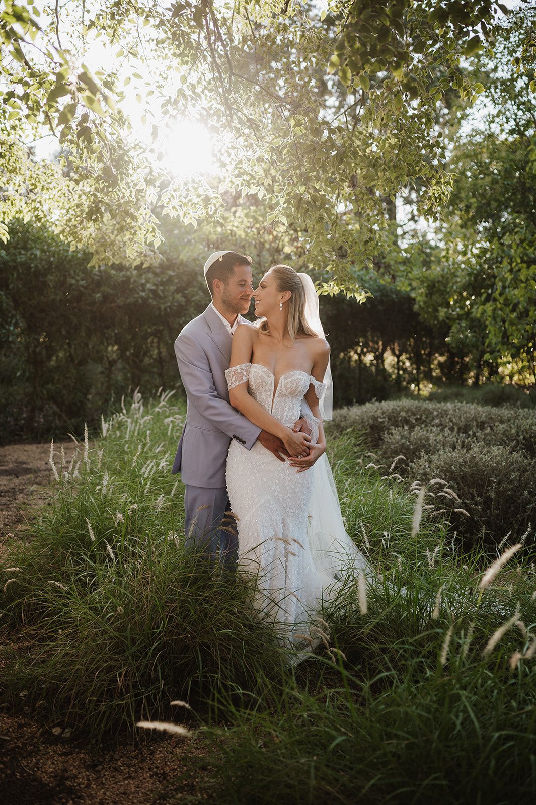 Bride and groom embracing in golden sunlight surrounded by lush green garden.