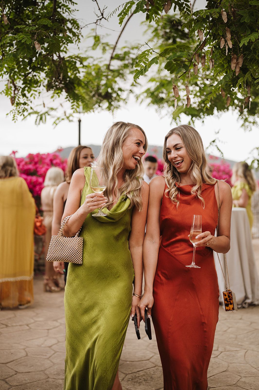 Two blonde women in green and rust satin dresses laughing and holding champagne at an outdoor wedding.