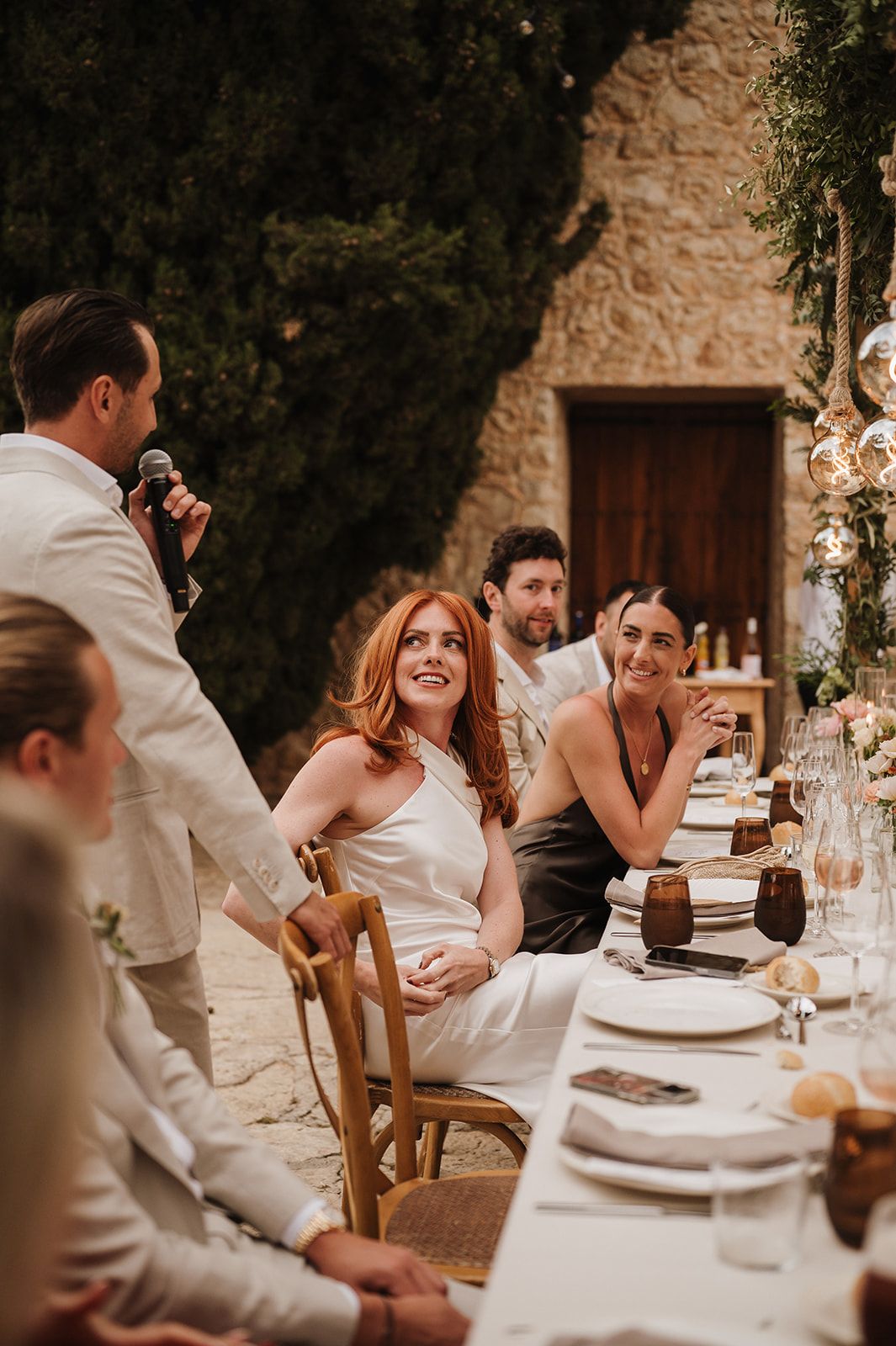 Bride with red hair laughs joyfully during wedding speech at outdoor stone venue reception dinner.