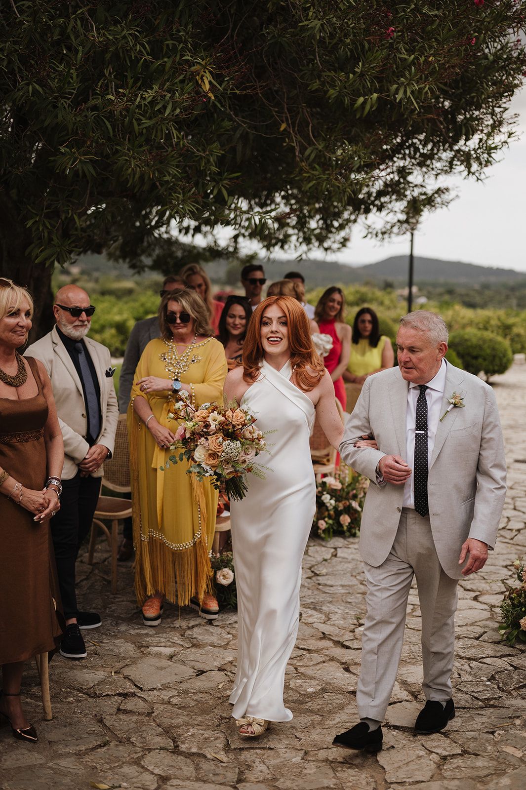 Radiant red-haired bride in white halter gown walks aisle with father at outdoor garden wedding.