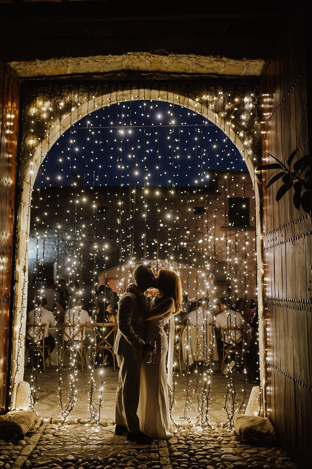 Bride and groom kissing under a glowing fairy light arch at night wedding venue.