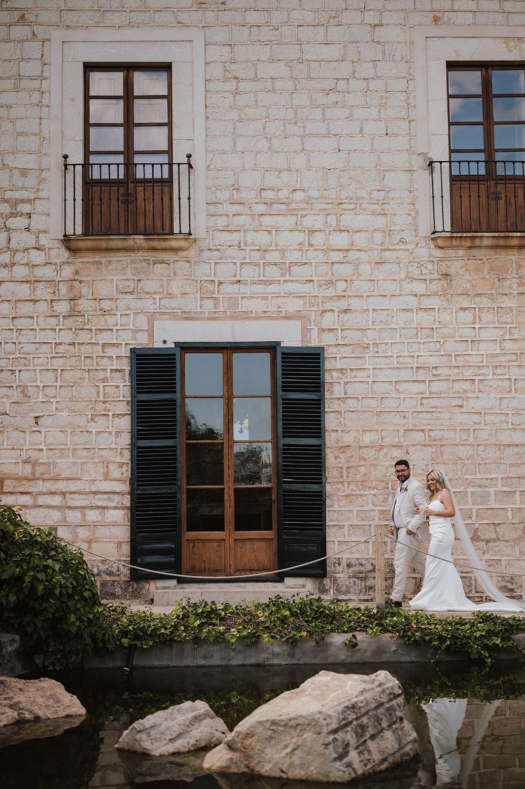 Bride and groom pose together in front of a rustic stone building with shuttered windows.