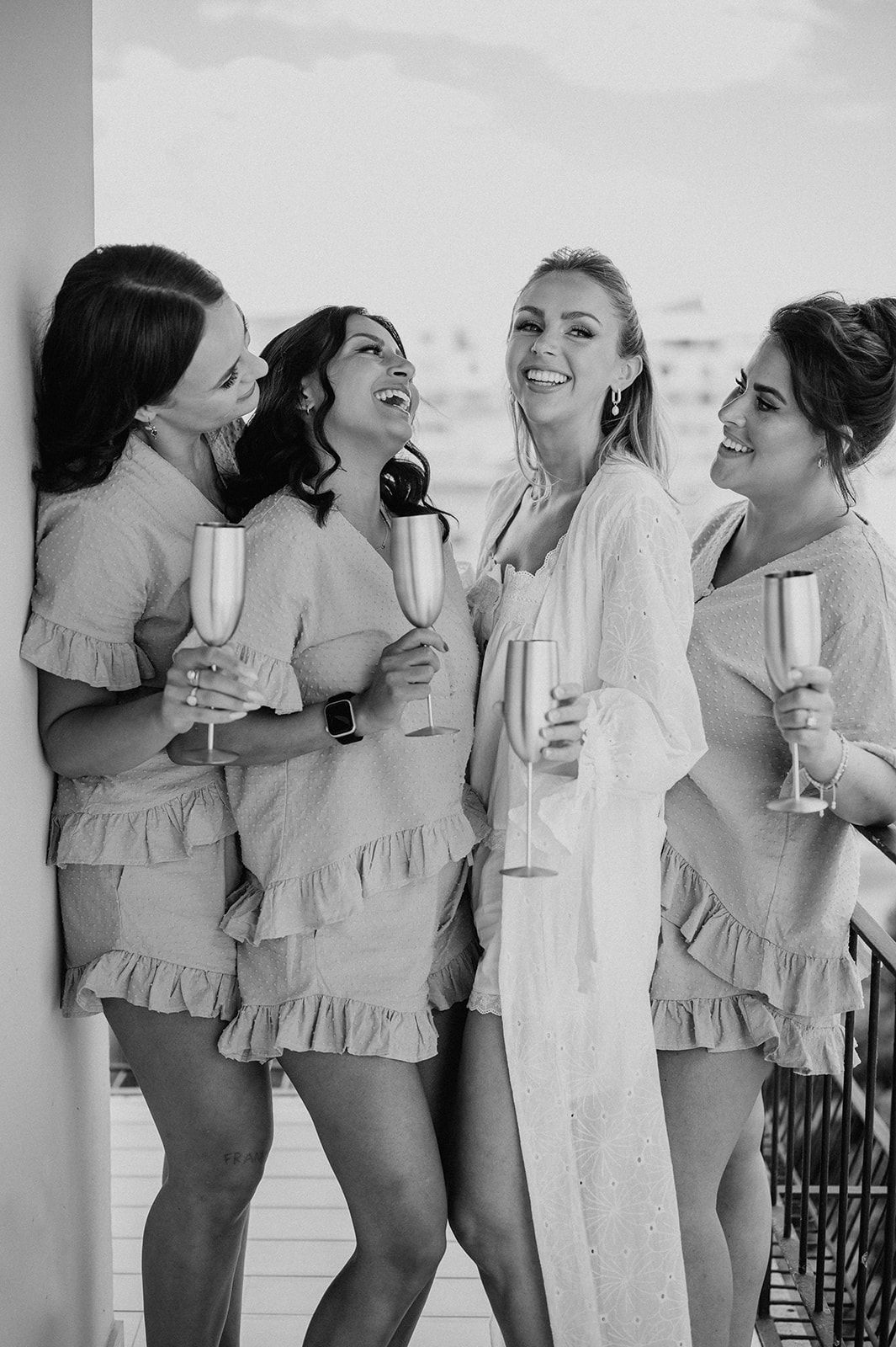 Four bridesmaids in matching ruffled sets laughing and toasting with champagne flutes on a balcony.