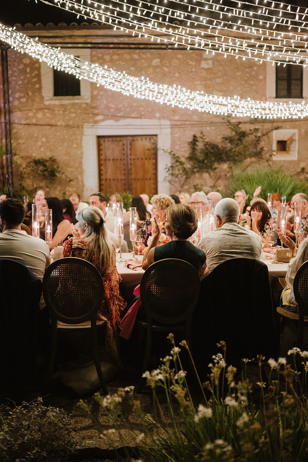 Wedding guests dining at candlelit outdoor reception table adorned with fairy lights at night.