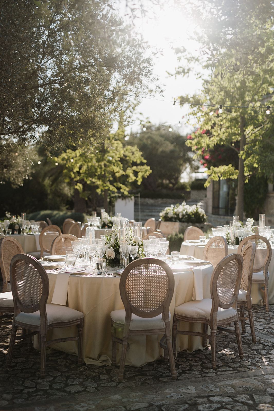 Elegant outdoor wedding reception with round tables, cane chairs, and floral centerpieces under trees.