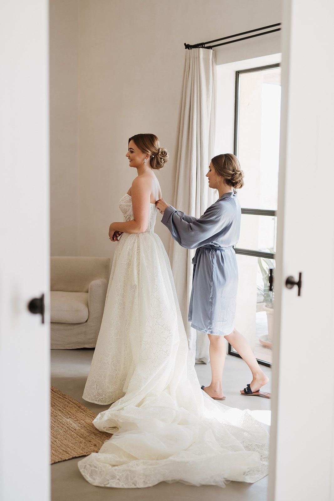 Bridesmaid in blue robe fastening the back of a bride's elegant lace wedding gown near a bright window.