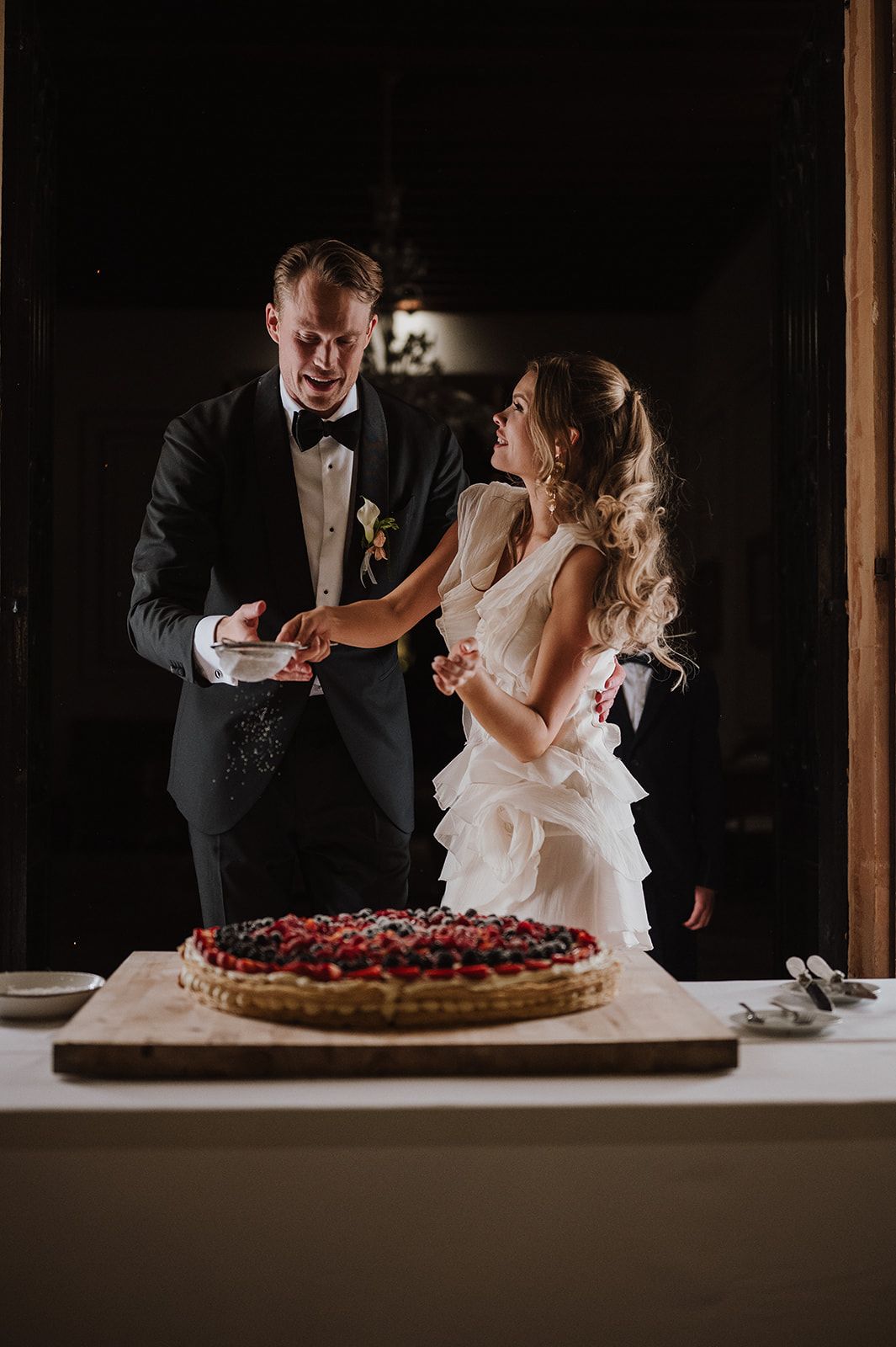 Bride and groom cut elegant berry tart wedding cake together at romantic dimly lit reception.
