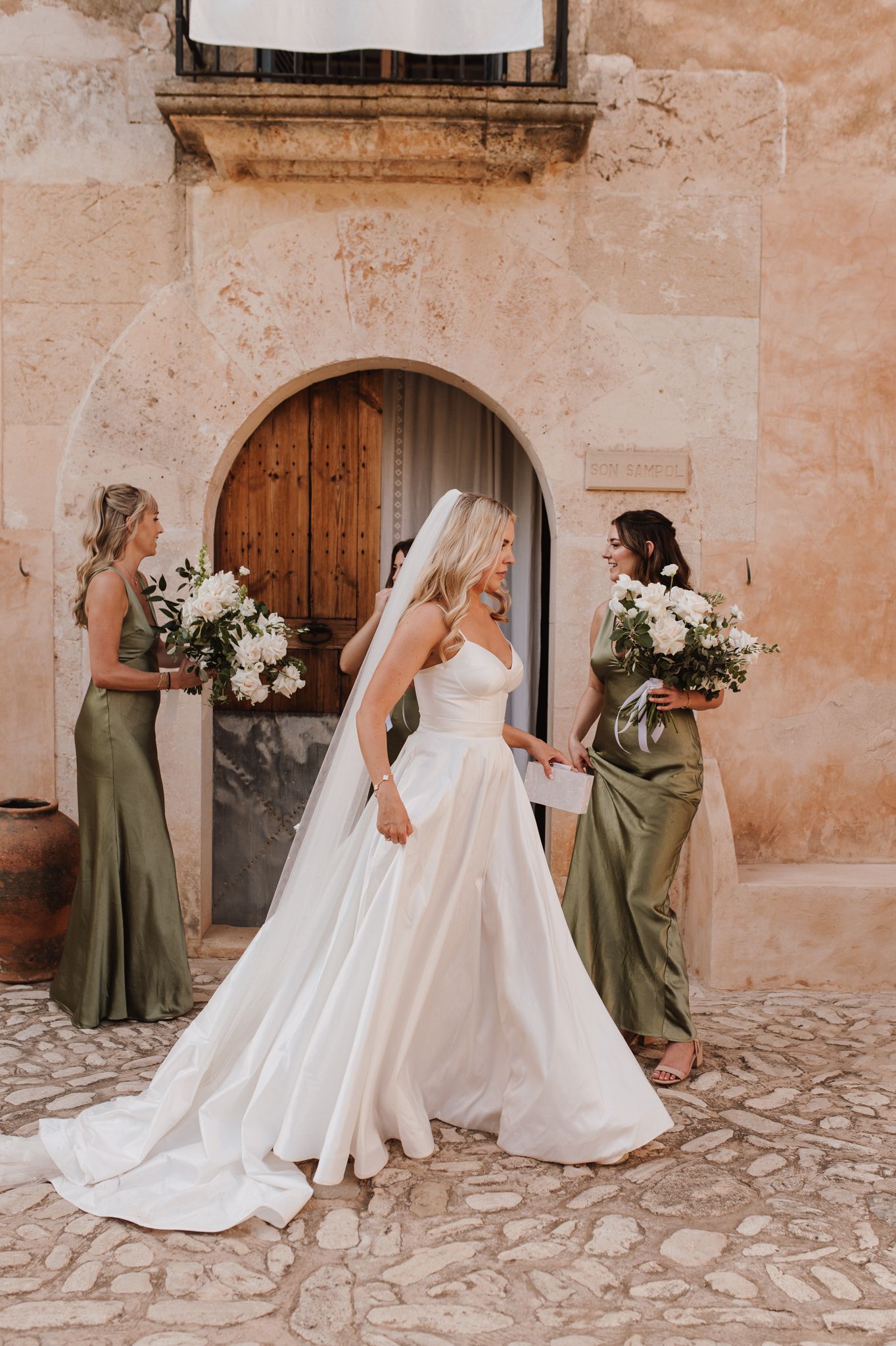 Bride in white gown stands with two bridesmaids in olive green dresses outside a rustic stone building.