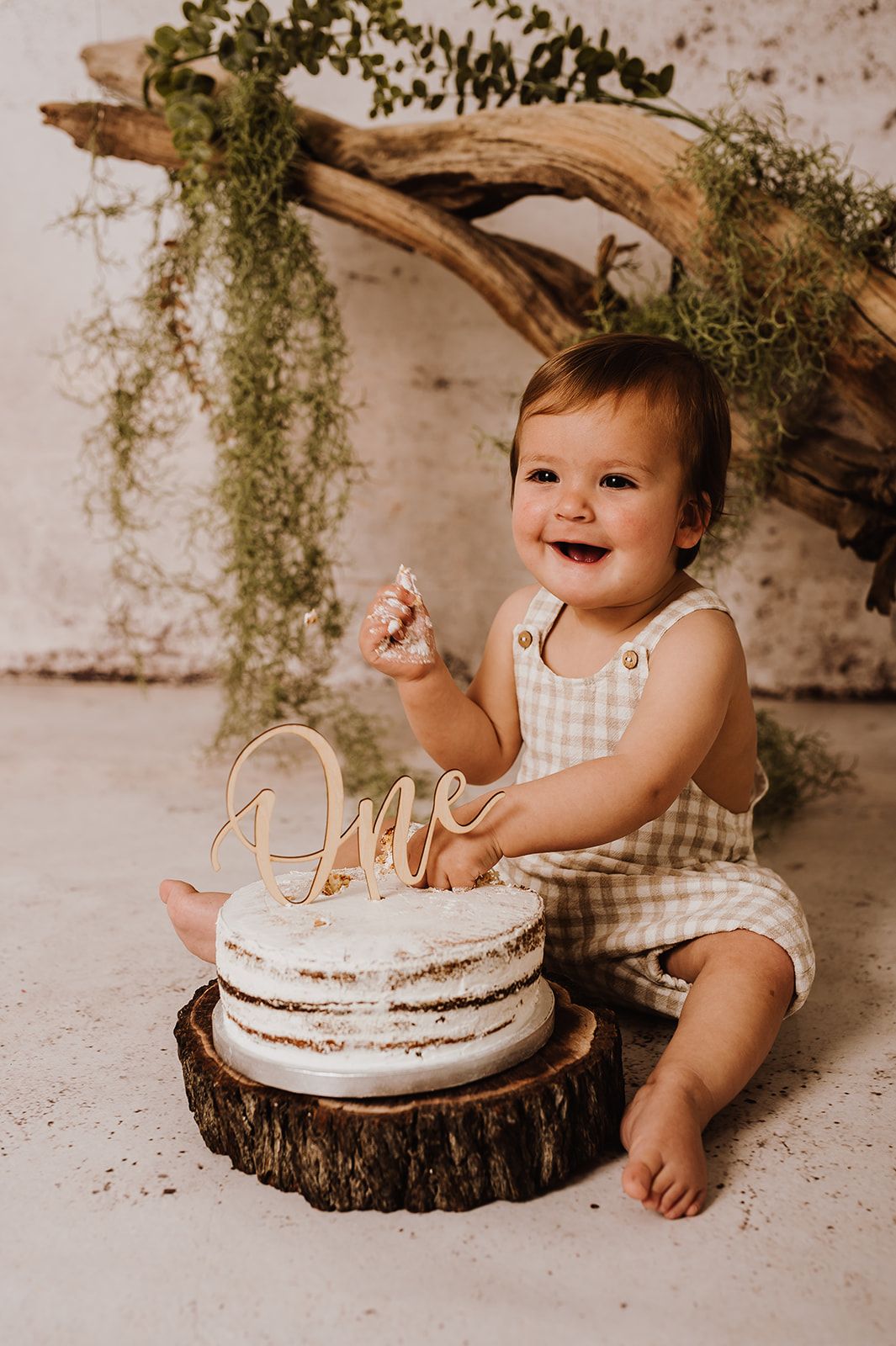 Smiling baby in gingham romper touching a naked cake with an One topper on a wood slice.