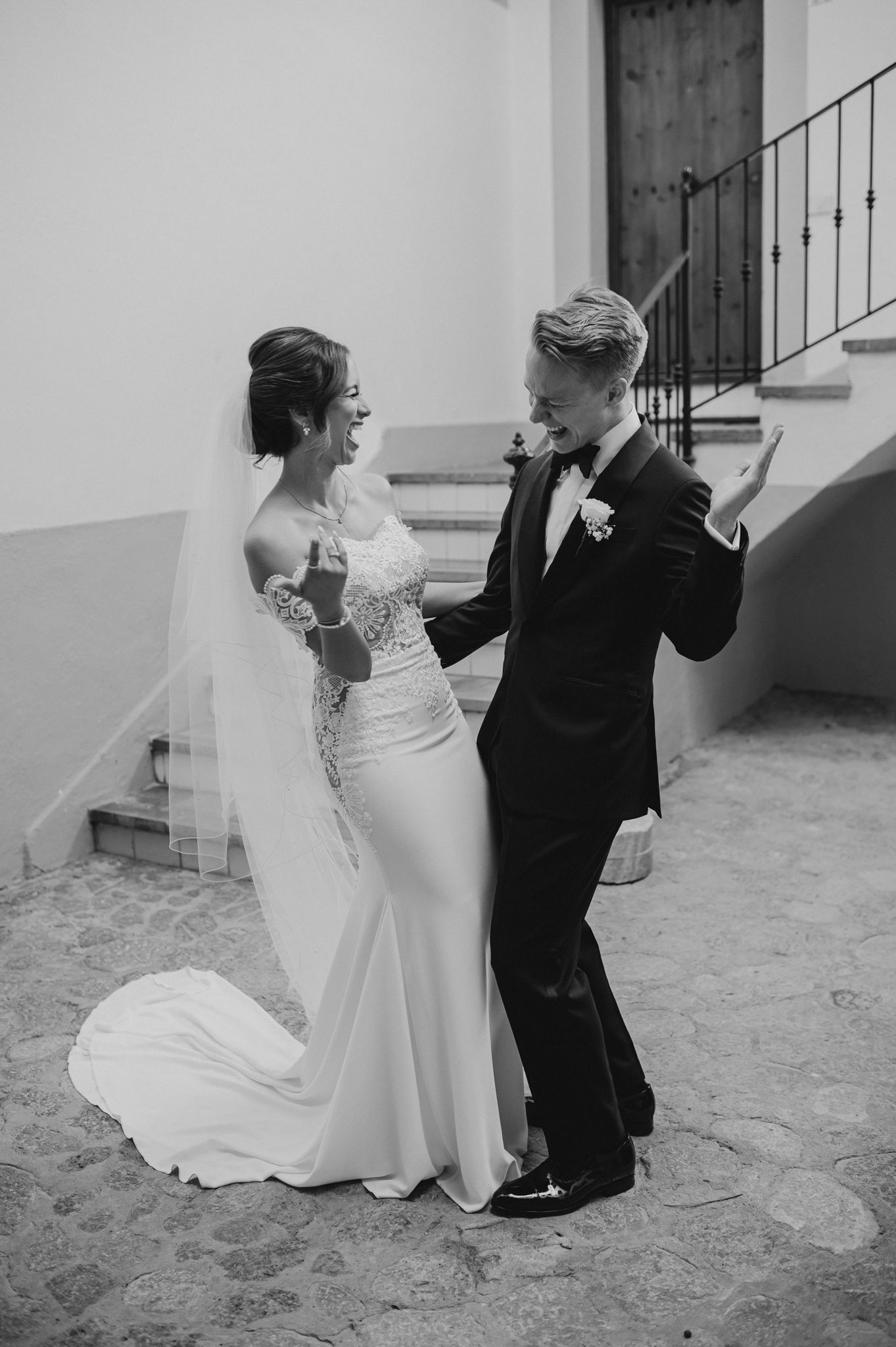 Black and white photo of a smiling bride in lace gown and groom in tuxedo dancing joyfully outside on their wedding day.