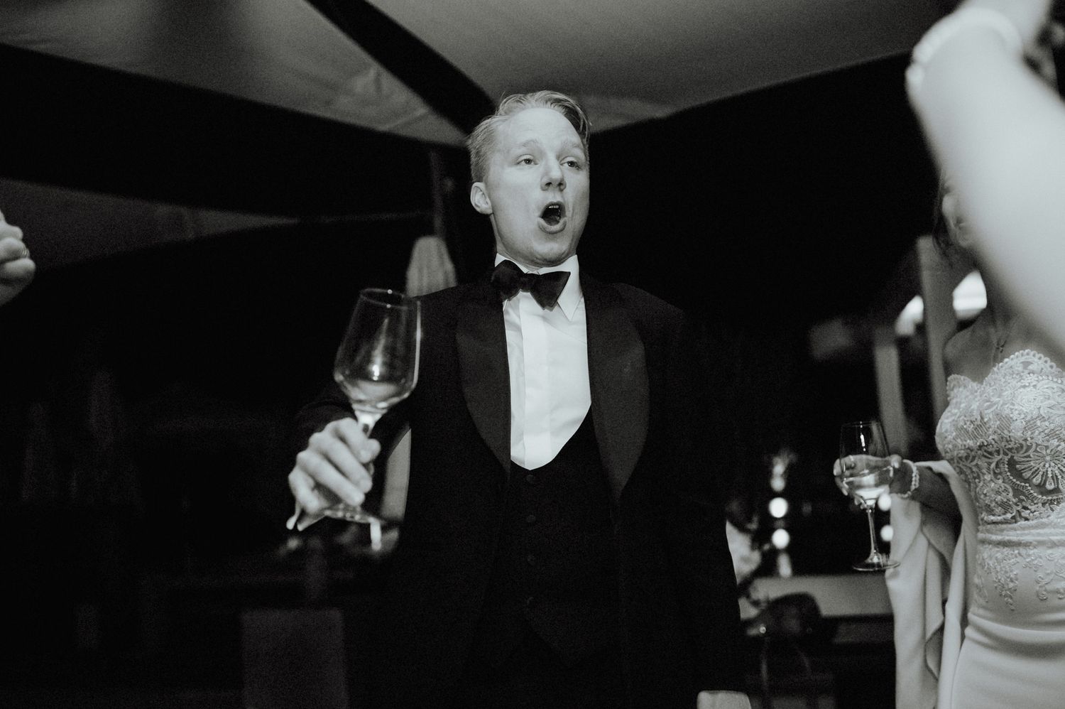 Excited groom in tuxedo raises a wine glass during wedding reception in black and white photo.