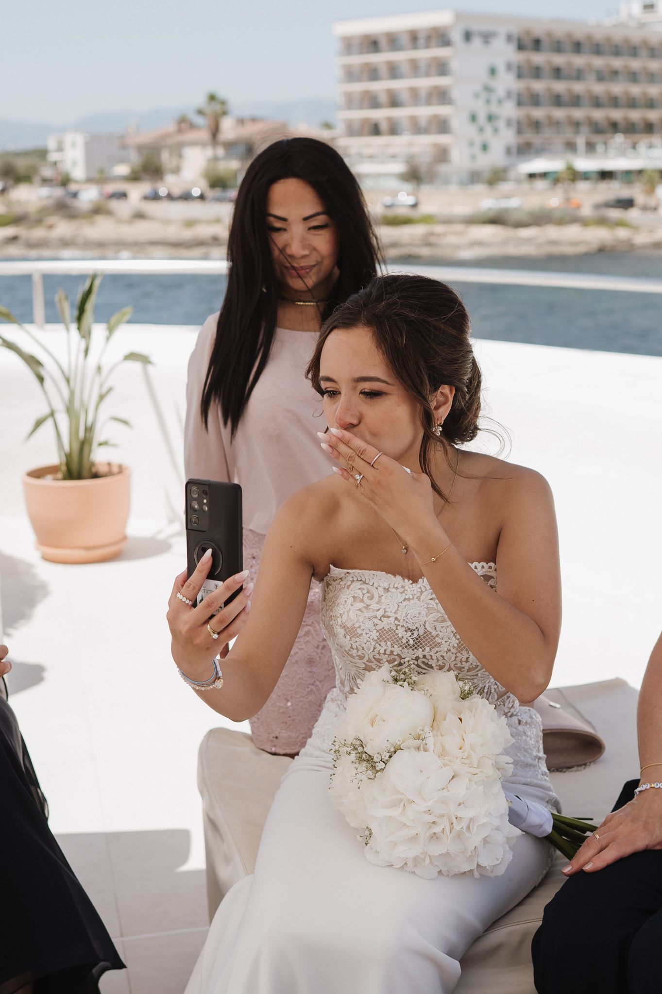 Emotional bride in strapless gown holds white bouquet while video calling during wedding ceremony.