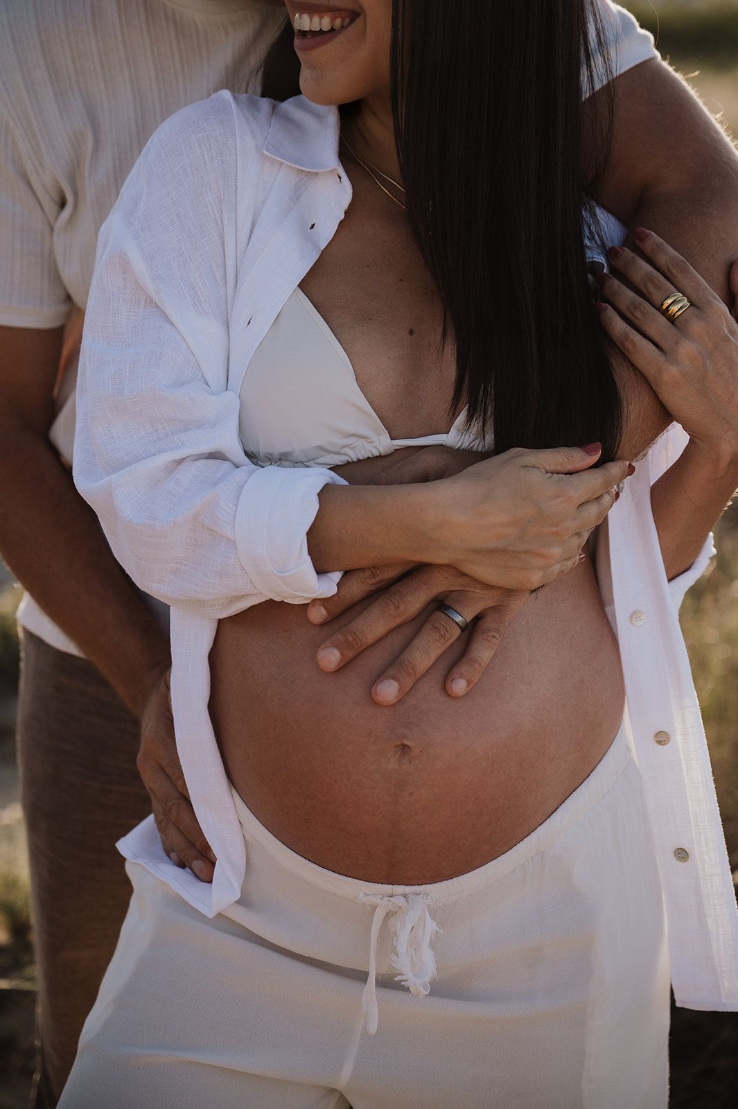 Close-up of pregnant womans bare baby bump being tenderly held by couple, woman wearing open white shirt.