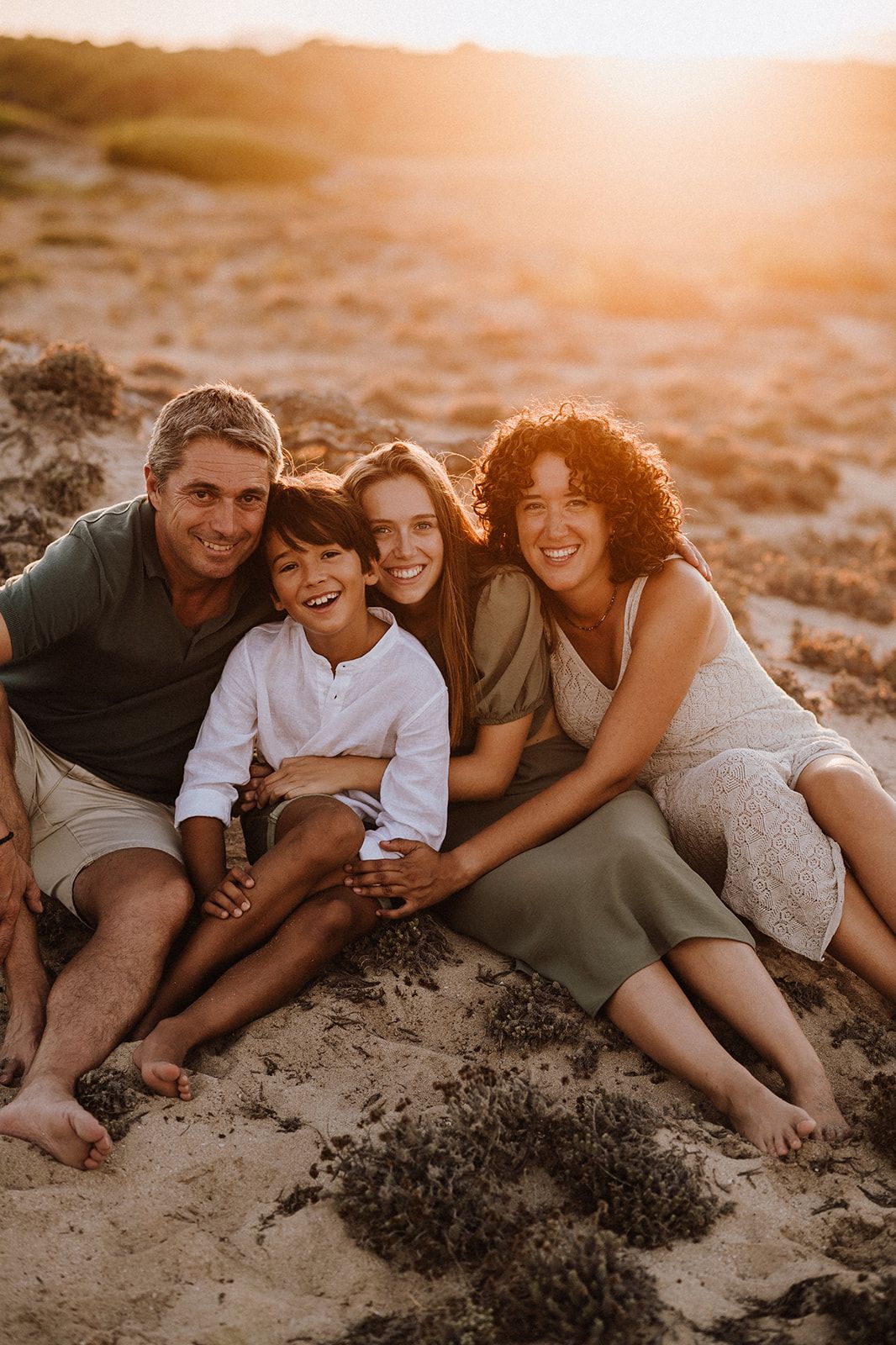 A happy family of four sits together on sandy dunes bathed in warm golden sunset light.
