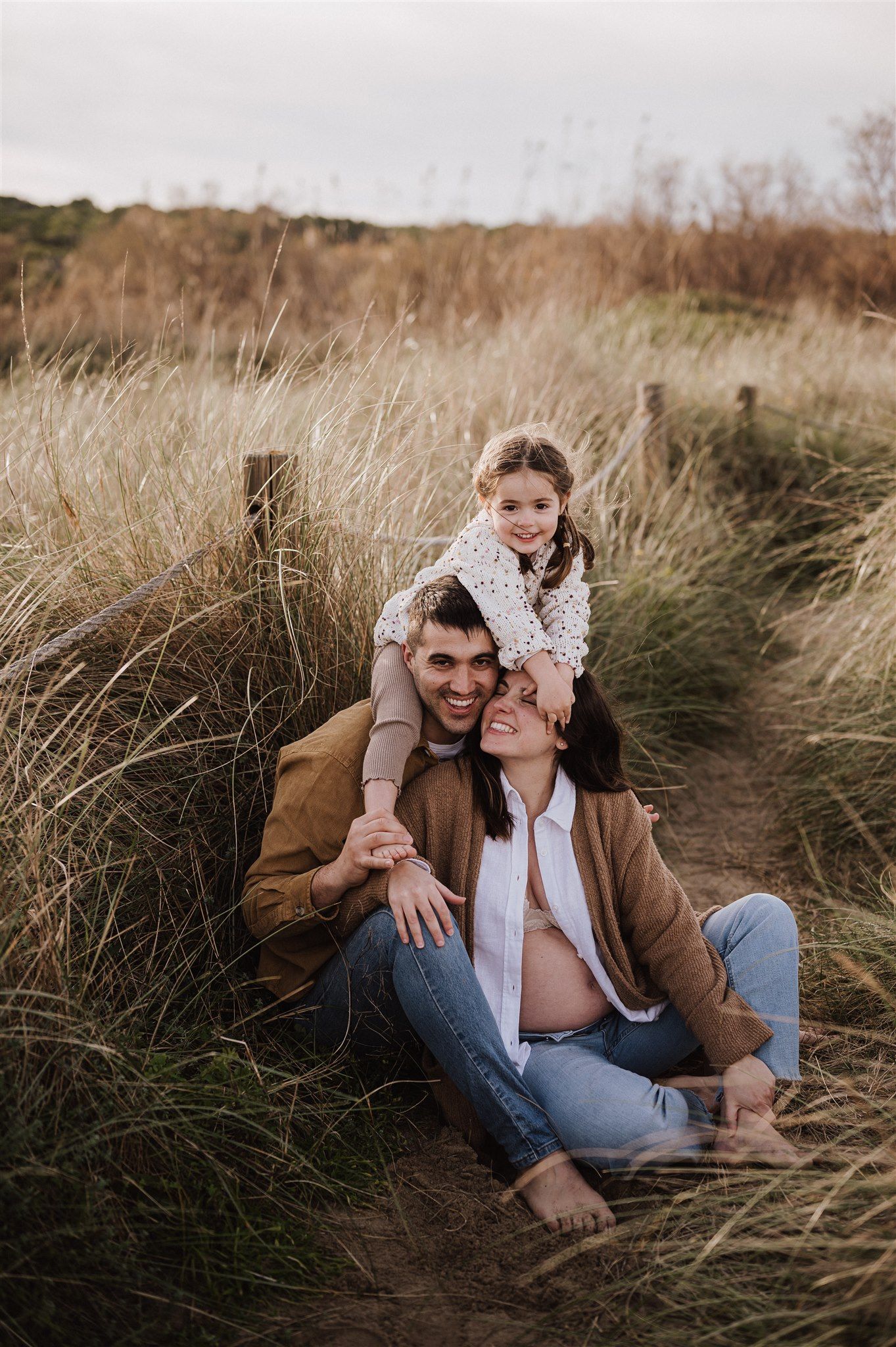 A happy family of three sits in tall grass dunes, with a young girl perched on her parents shoulders outdoors.