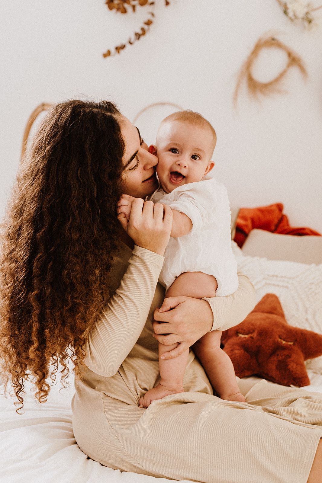 Curly-haired mother kissing her laughing baby while sitting on a white bed with boho decor.