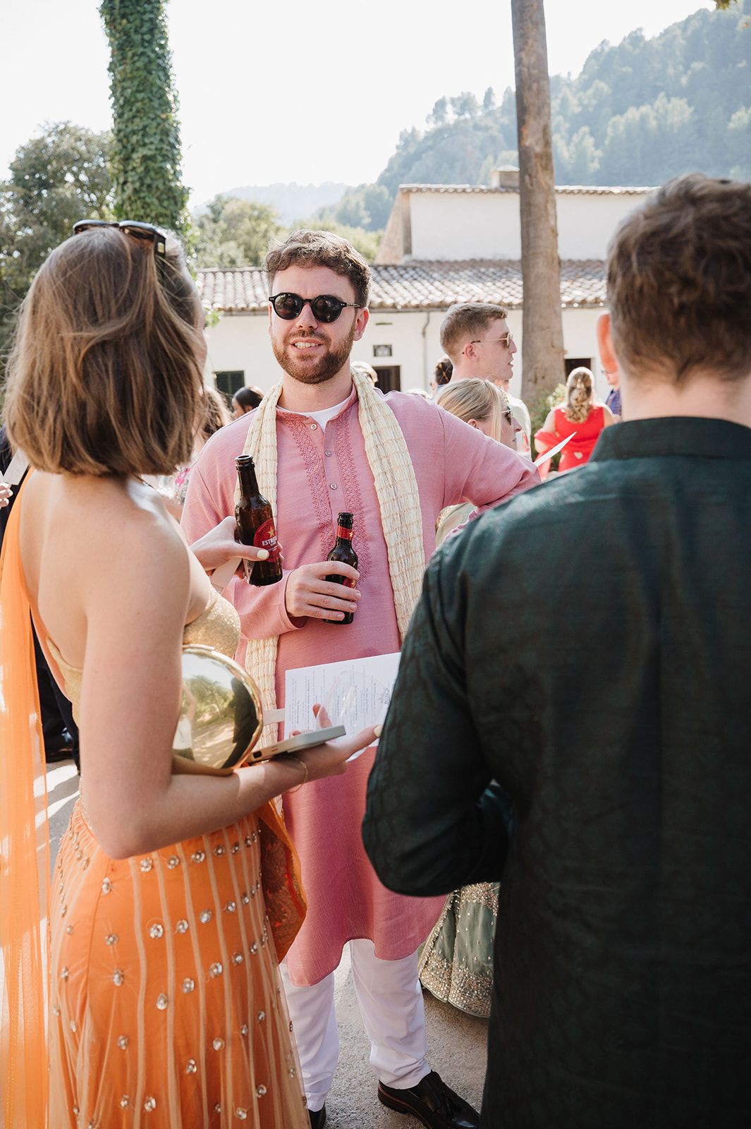 Male wedding guest in pink kurta and sunglasses holding beer bottle at outdoor Indian wedding reception.