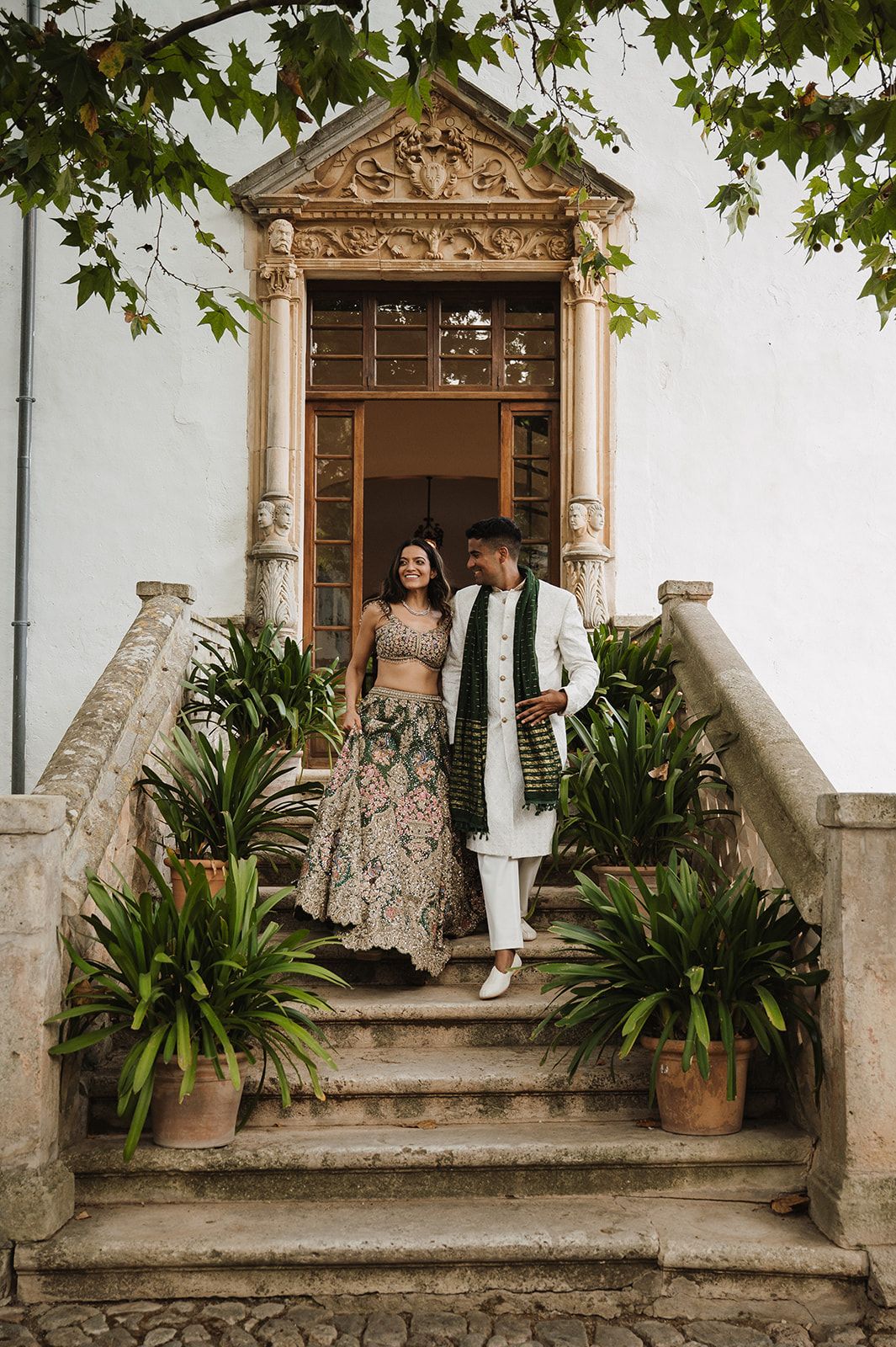 Indian bride and groom in traditional attire descend ornate stone steps of a white villa entrance.