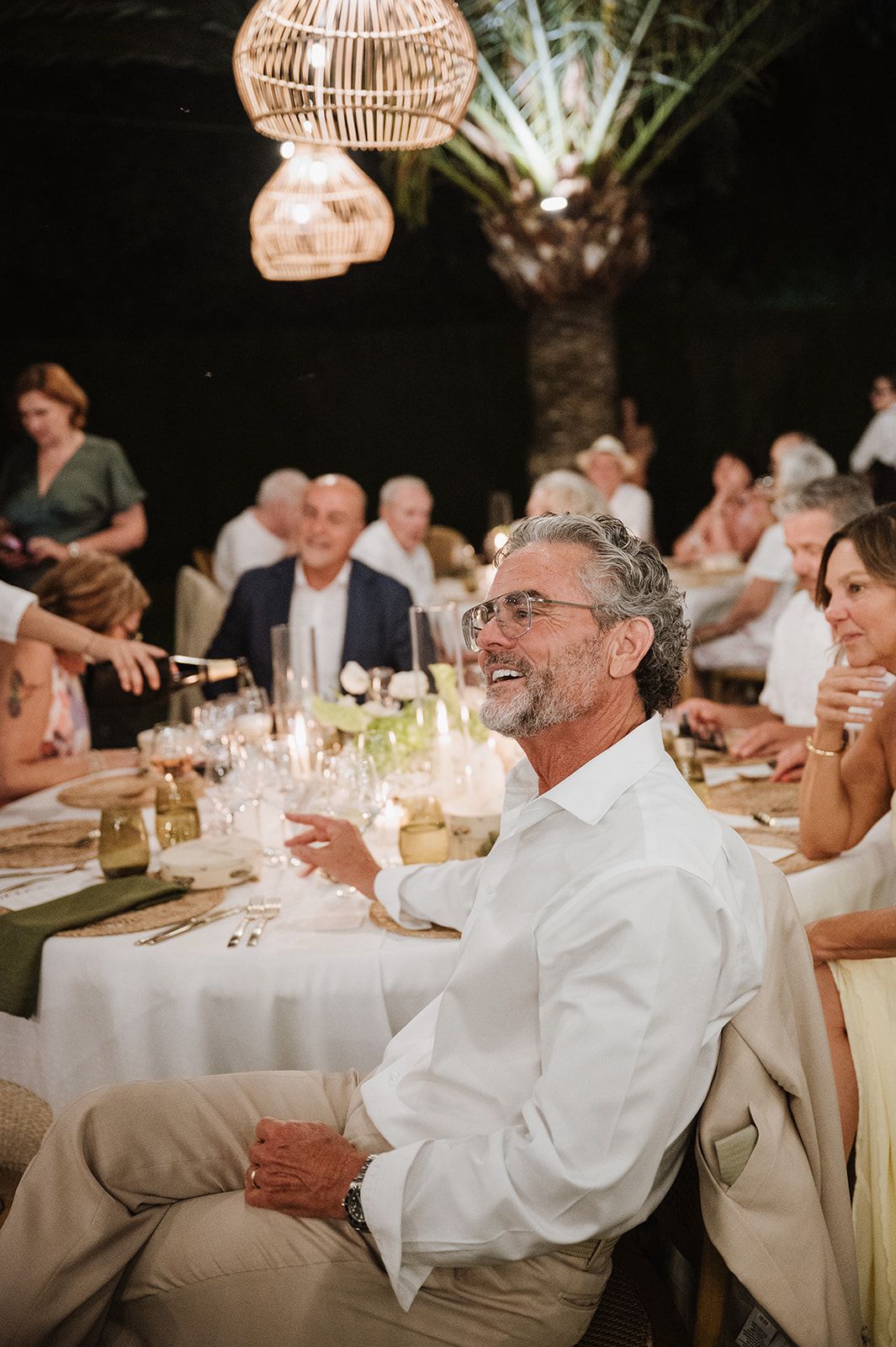 Smiling bearded man in white shirt enjoys conversation at candlelit outdoor wedding reception under rattan pendant lights.