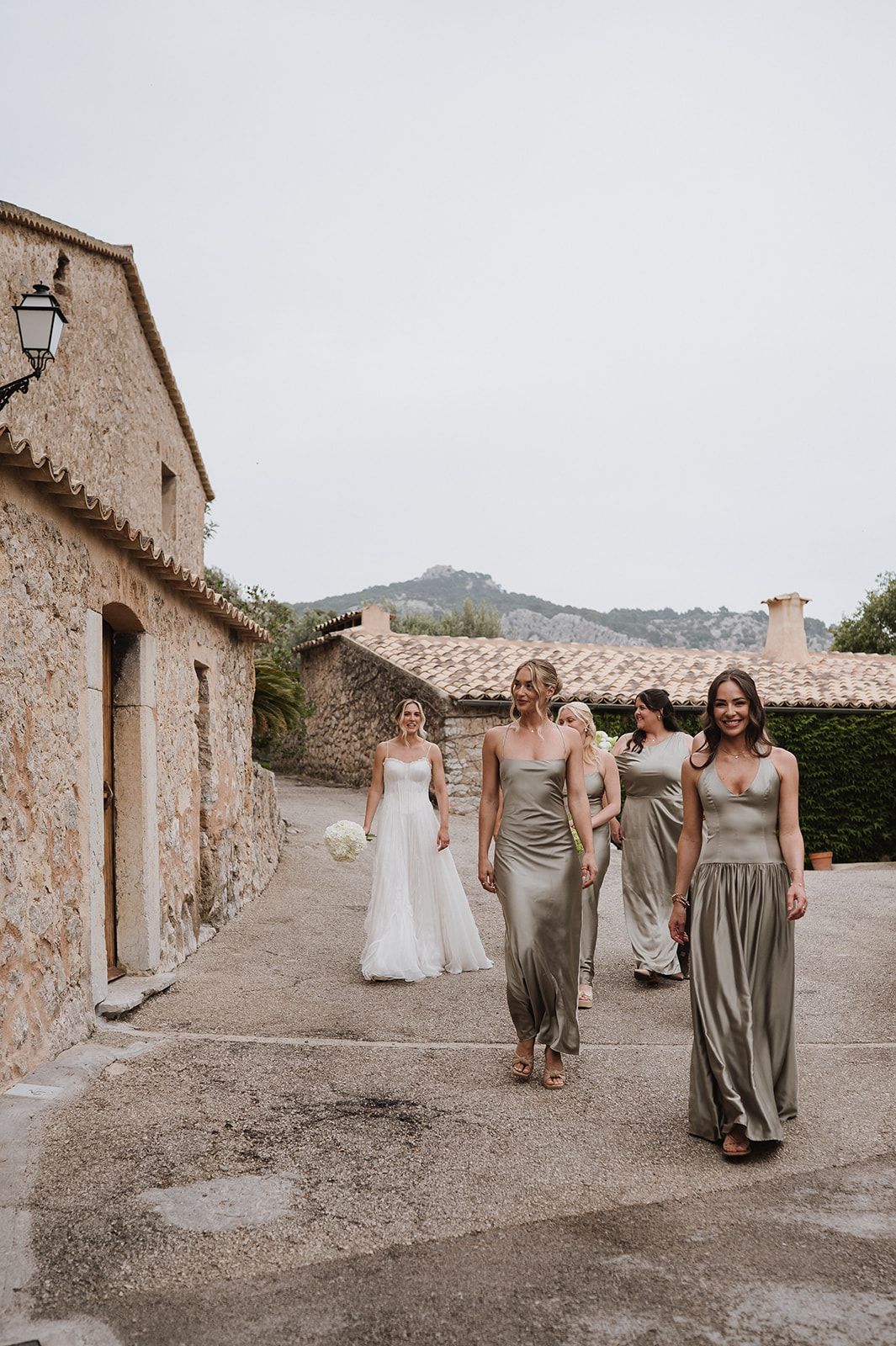 Bride in white gown walks with bridesmaids in sage green dresses at rustic stone venue.