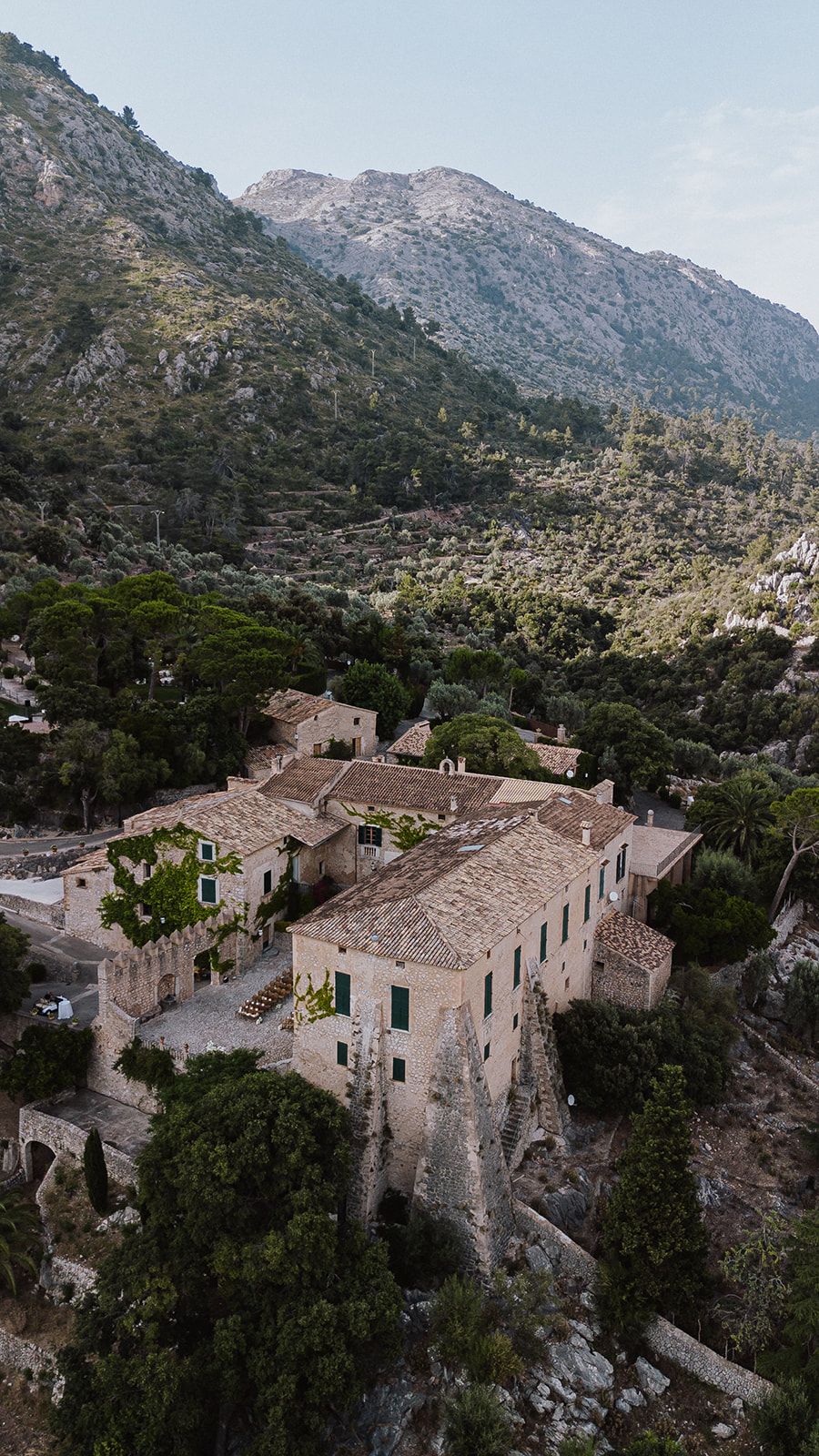 Aerial drone view of historic stone estate nestled among lush green mountains in Mediterranean landscape.