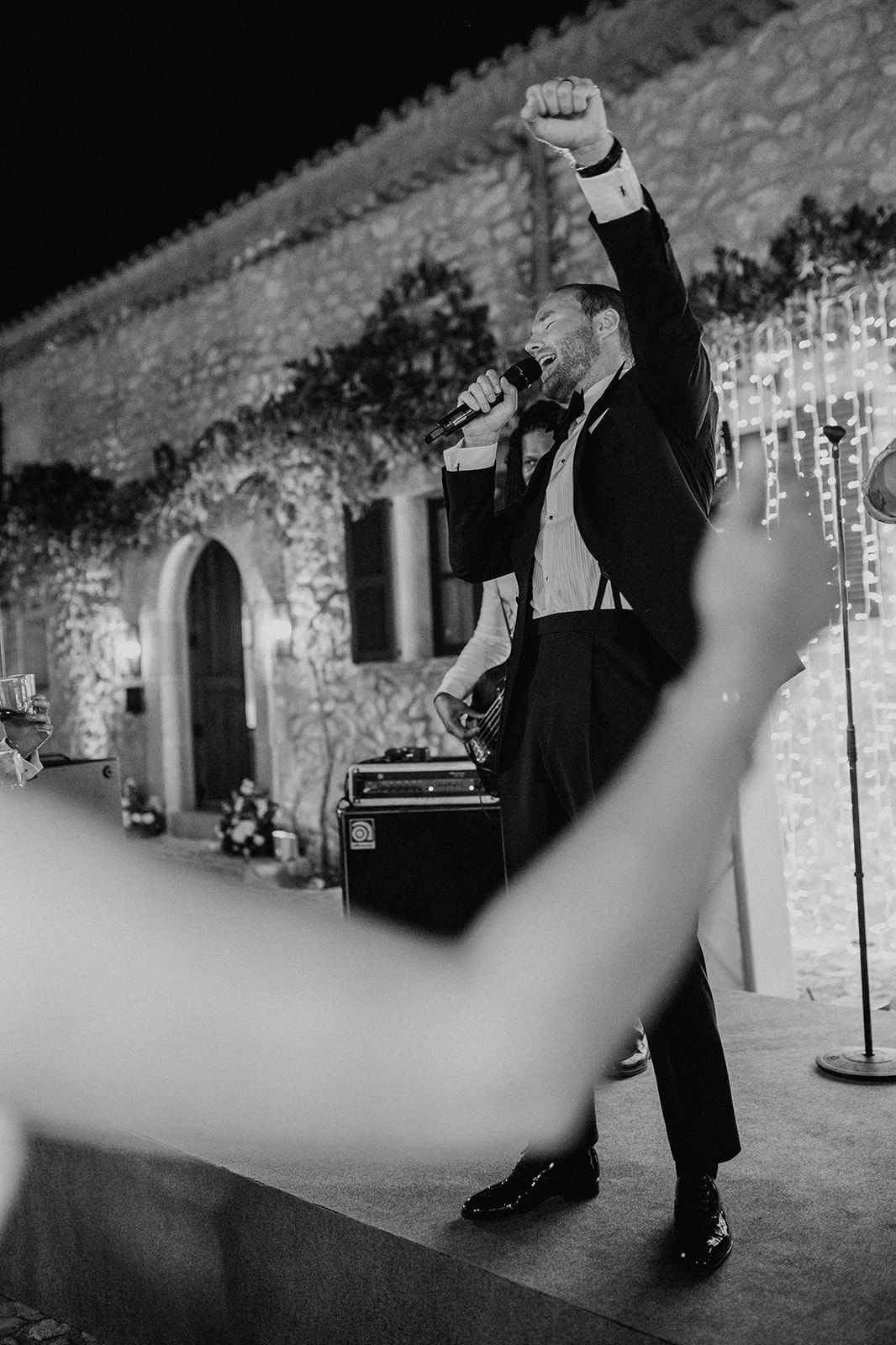 Energetic wedding performer in tuxedo raises fist while singing on stage at night reception.