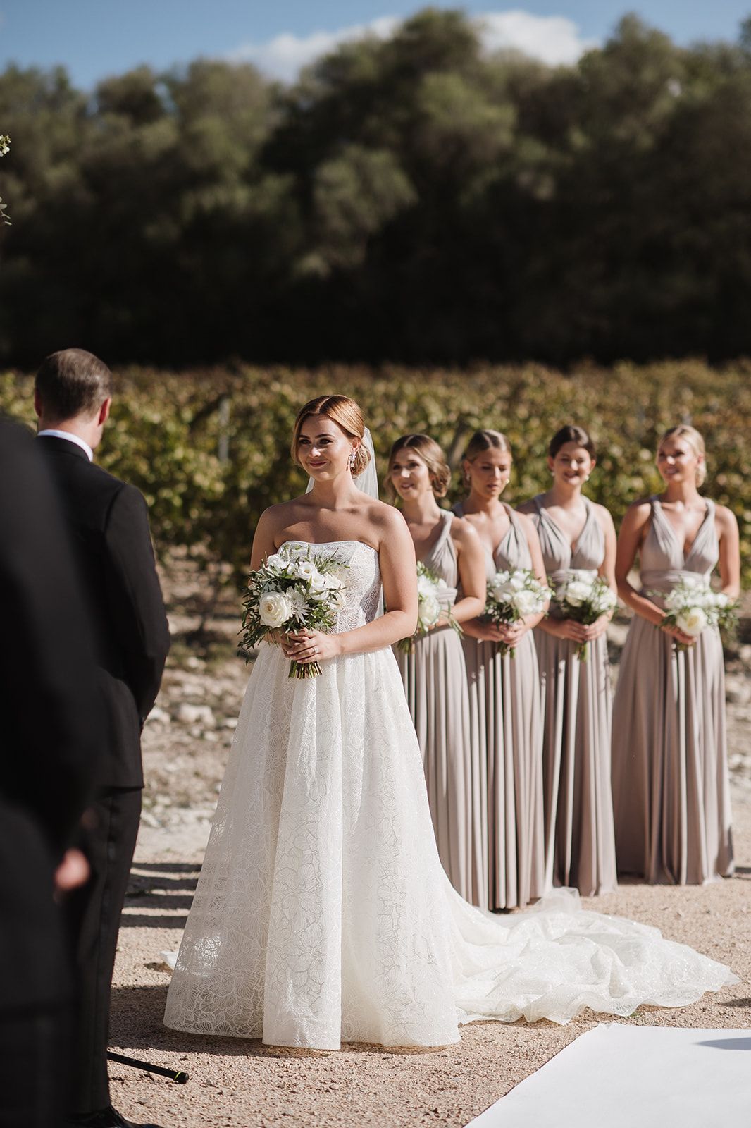 Bride in strapless lace gown at outdoor vineyard ceremony with bridesmaids in taupe dresses.