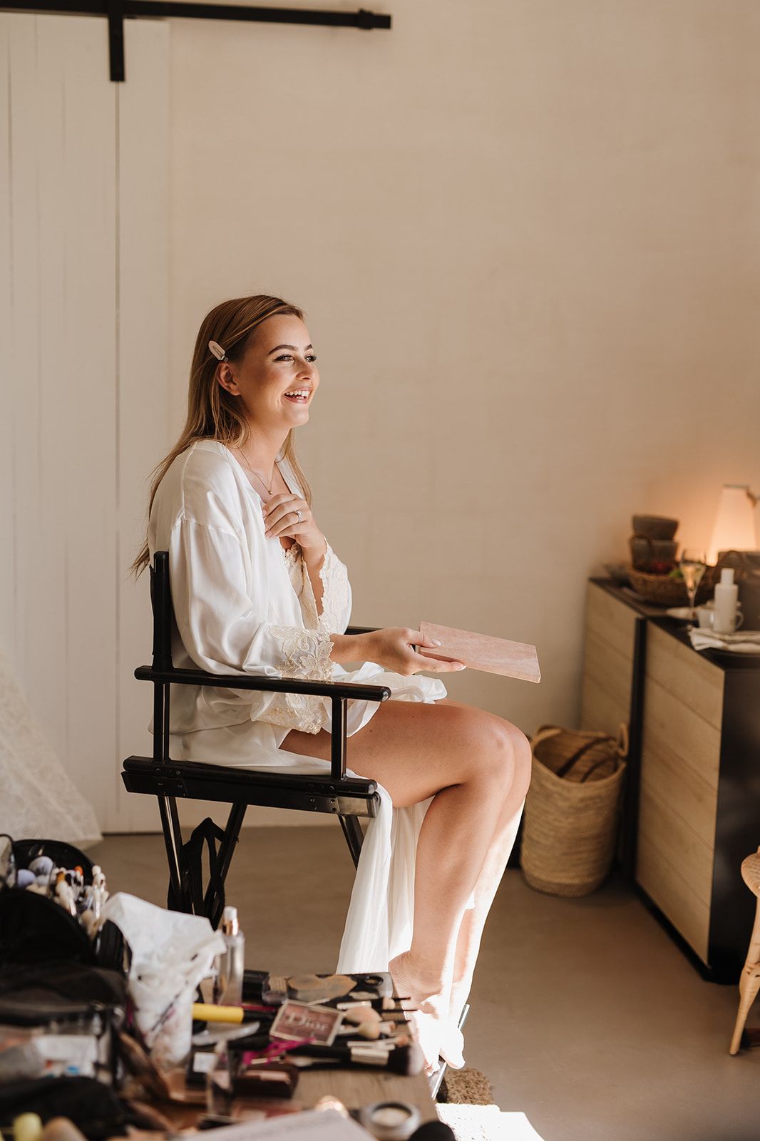Smiling bride in white robe sitting in makeup chair holding a gift box in a bright bridal suite.